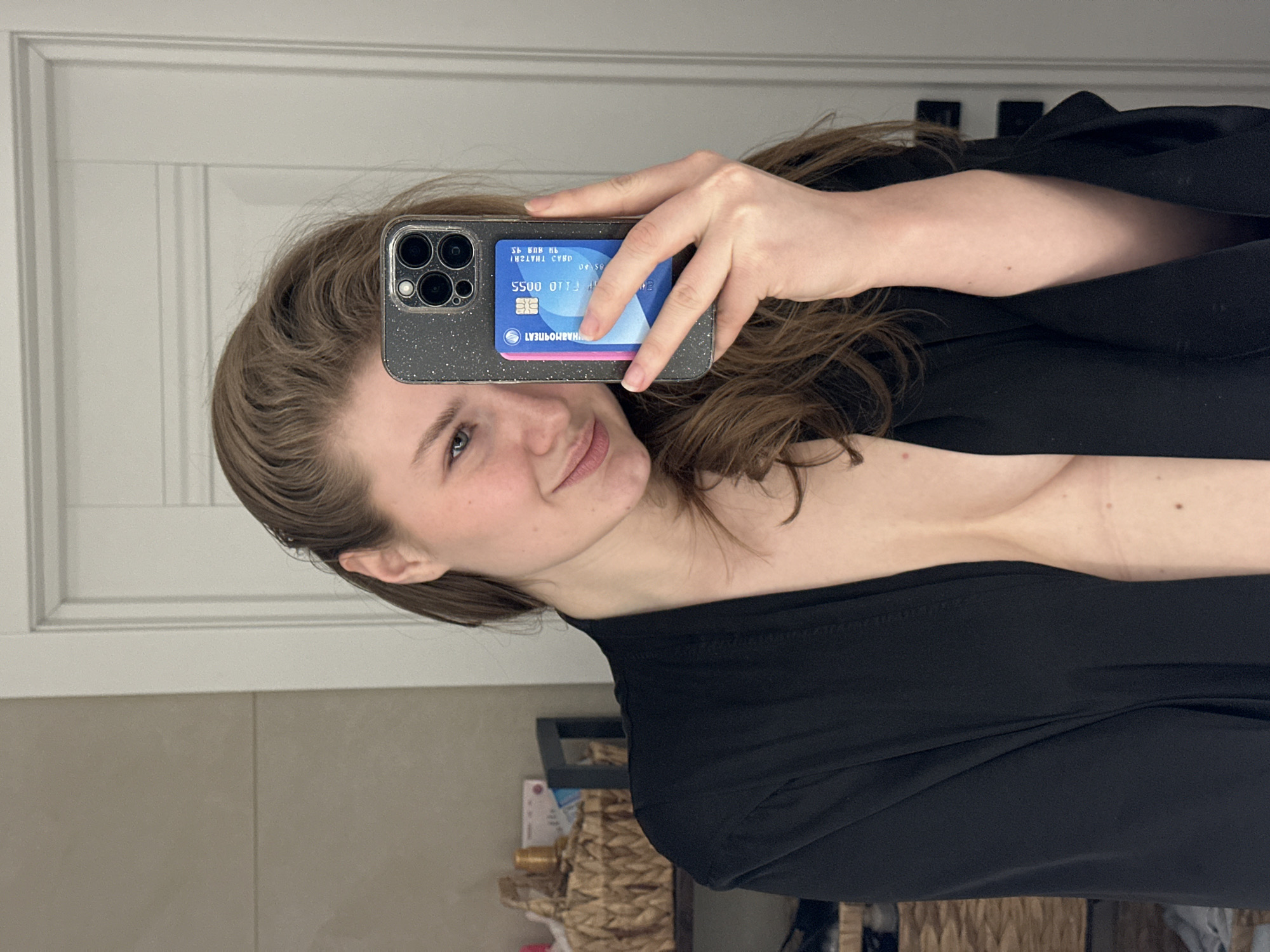 The image displays a female individual taking a mirror selfie indoors. The setting is a private residence or hotel room, inferred to be in Kazan, Tatarstan, Russia, based on the visible "МАРМОРИТБАНК" (Marmoritbank) and "ТАТАРСТАН" (Tatarstan) text on a credit card.
The subject is a fair-skinned female with light brown hair, depicted from the chest upwards. She wears an open black garment, possibly a robe or kimono, revealing her chest. She exhibits a slight smile.
In her right hand, she holds a dark grey smartphone, encased in a glittery cover and featuring a triple-lens camera system. A blue credit card is partially visible within a cardholder attached to the phone's back. The card displays the reversed text "МАРМОРИТБАНК", "ТАТАРСТАН", and the partial number "5500 0710 0055".
The background on the left consists of a light-colored tiled wall or surface. A woven basket containing various bottles and containers is situated on a shelf, alongside a dark rectangular frame. The background on the right reveals a white paneled door with horizontal molding details and a dark door handle mechanism.