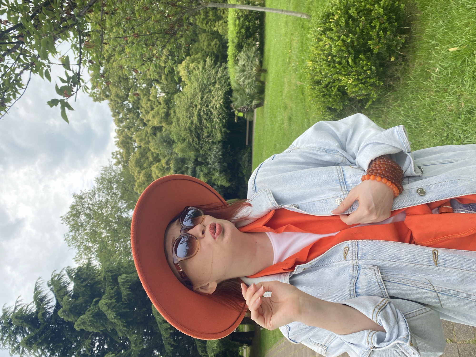 A woman is photographed in a park setting. She is wearing a wide-brimmed hat, sunglasses, an orange top, and a denim jacket. She has a beaded bracelet on her wrist and is holding the edge of her jacket. The background consists of trees, shrubs, and green grass. The sky is cloudy. The location is Sochi, Russia.