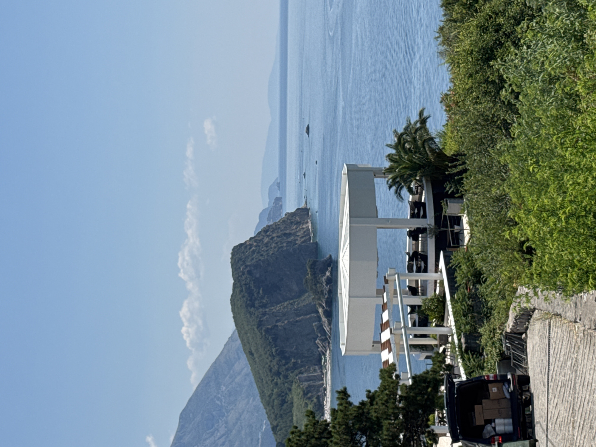 The image depicts a scenic view of the Adriatic Sea near the city of Budva, Montenegro. In the foreground, a black van with its rear hatch open is parked on a paved pathway. Several cardboard boxes and cylindrical containers are visible inside the van, suggesting a delivery or transport operation. To the right of the van, a pile of metal frames leans against a stone wall, surrounded by lush green foliage.
A modern, white-roofed structure with an open-air seating area and tables is situated on an elevated terrace overlooking the sea. A palm tree stands next to the structure. The middle ground is dominated by the vast blue expanse of the sea, with a large, verdant, rocky island prominent in the distance. Further beyond, mountainous terrain and a hazy coastline complete the horizon under a clear blue sky with scattered white clouds. The overall scene is one of a coastal resort area on a bright, sunny day.