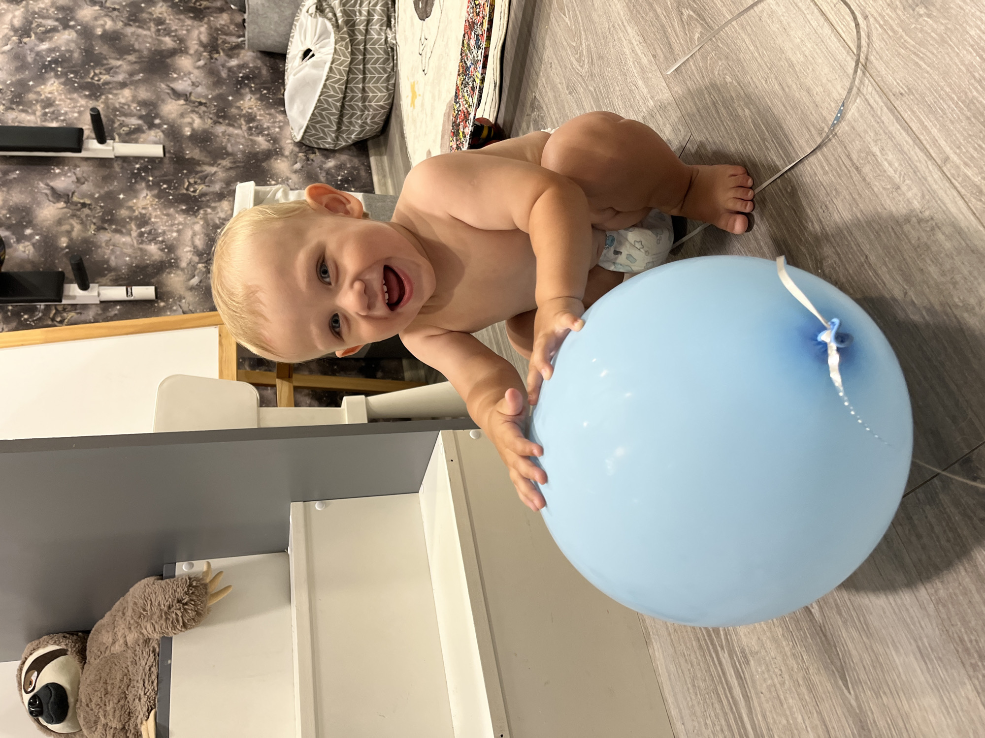A young child with blonde hair and blue eyes is playing on the floor with a blue balloon. The child is on their hands and knees, looking up at the camera with a wide smile and open mouth. They are wearing a diaper. The floor is made of light-colored wood planks. In the background, there is a grey upholstered laundry basket with a geometric pattern, a child's play mat with colorful designs, and part of a baby changing table or dresser. A stuffed sloth toy is hanging from the edge of a grey shelf. The overall scene appears to be an indoor play area or bedroom. The lighting suggests it is daytime.