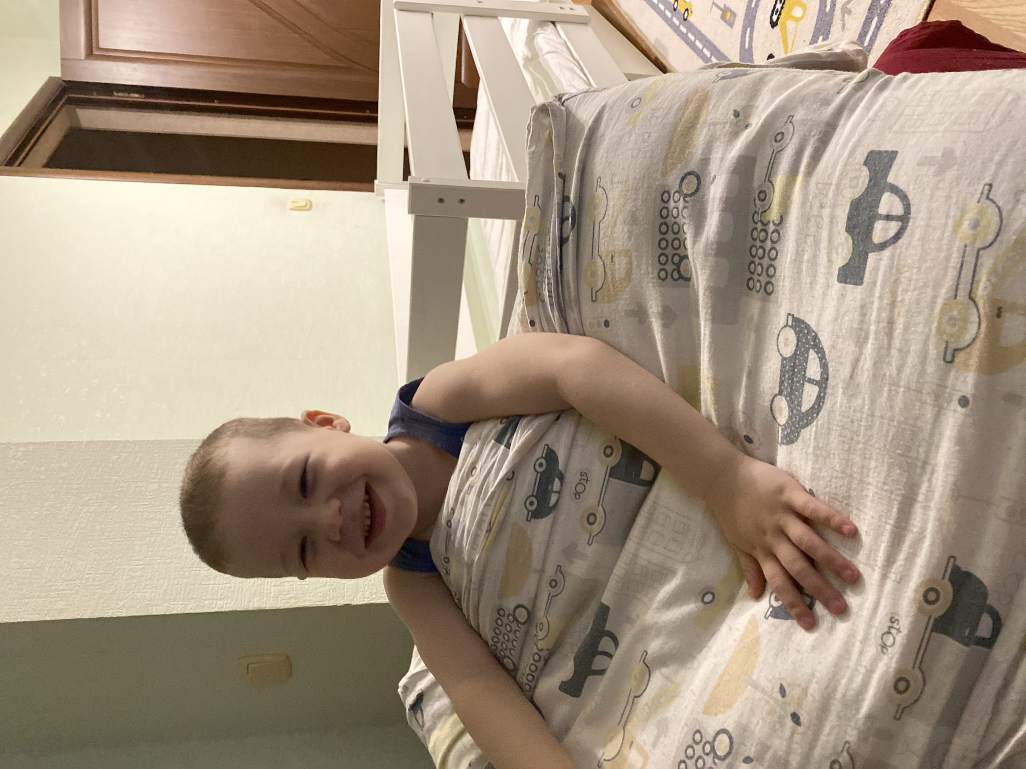 A child is lying on a bed covered with a car-themed blanket. The bed appears to be a top bunk, based on the visible frame. A wooden cabinet is partially visible in the background. The room has light green walls. The location is not ascertainable from the image, and there is no indication that it is in Sochi, Russia.