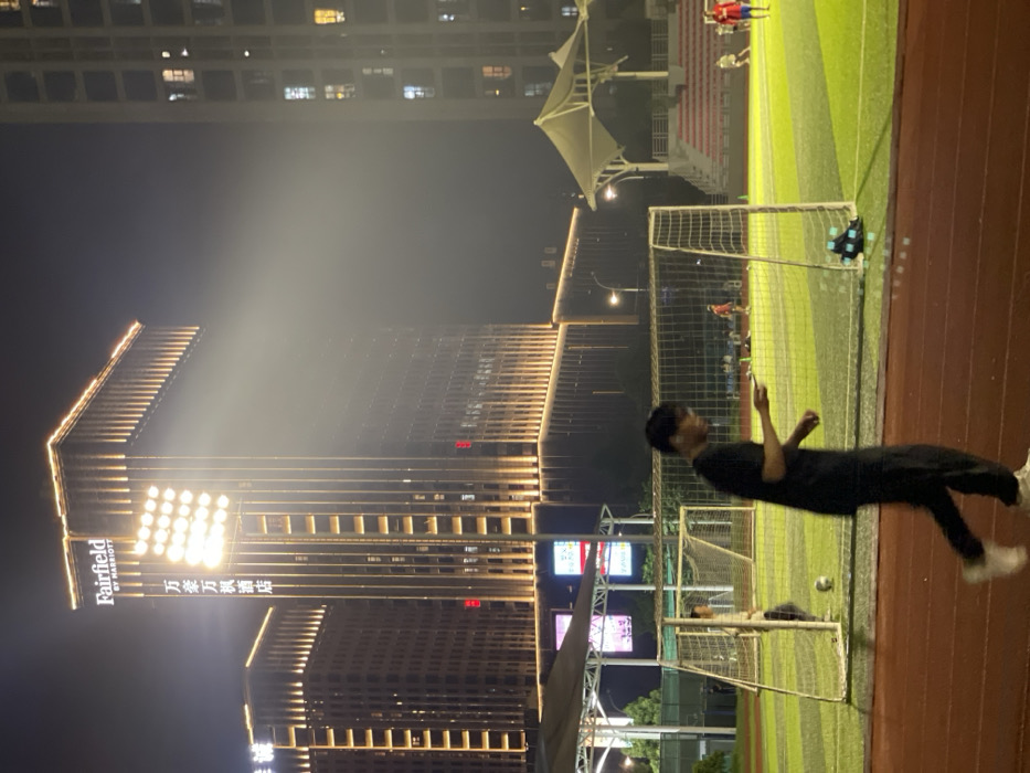 The image shows a man walking on a red running track at night. He is wearing a black shirt and black pants. He is looking down at his phone. In the background, there is a soccer field, soccer goal, and a large building with the sign “Fairfield by Marriott” illuminated with bright white lights. There are also people playing soccer on the field. The coordinates 30.177, 120.143 are in the city of Hangzhou, China. The scene is likely a sports complex within the city. The image was taken at night. The lighting is bright and the background is slightly hazy.