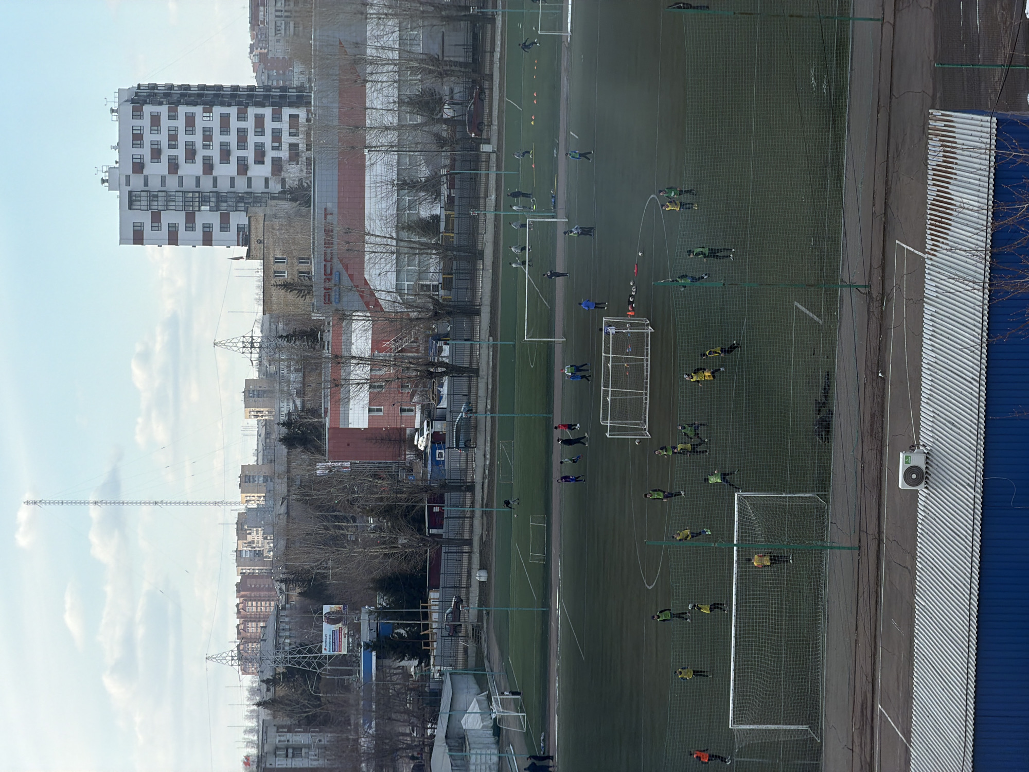 A high-angle, daytime view shows a soccer field in Krasnoyarsk, Russia, where a soccer practice is in progress. The field is rectangular and green, with white lines marking the boundaries. There are several soccer goals placed along the sidelines. Around 20-30 people are on the field, likely soccer players and coaches, wearing various colors of sportswear. Some are actively playing soccer, while others appear to be practicing drills. A tall white and gray residential building stands in the background to the left, alongside other urban structures and bare trees. A tall antenna is also visible against a light blue sky with scattered clouds. Adjacent to the soccer field is a gray pavement area and a building with blue corrugated metal siding.