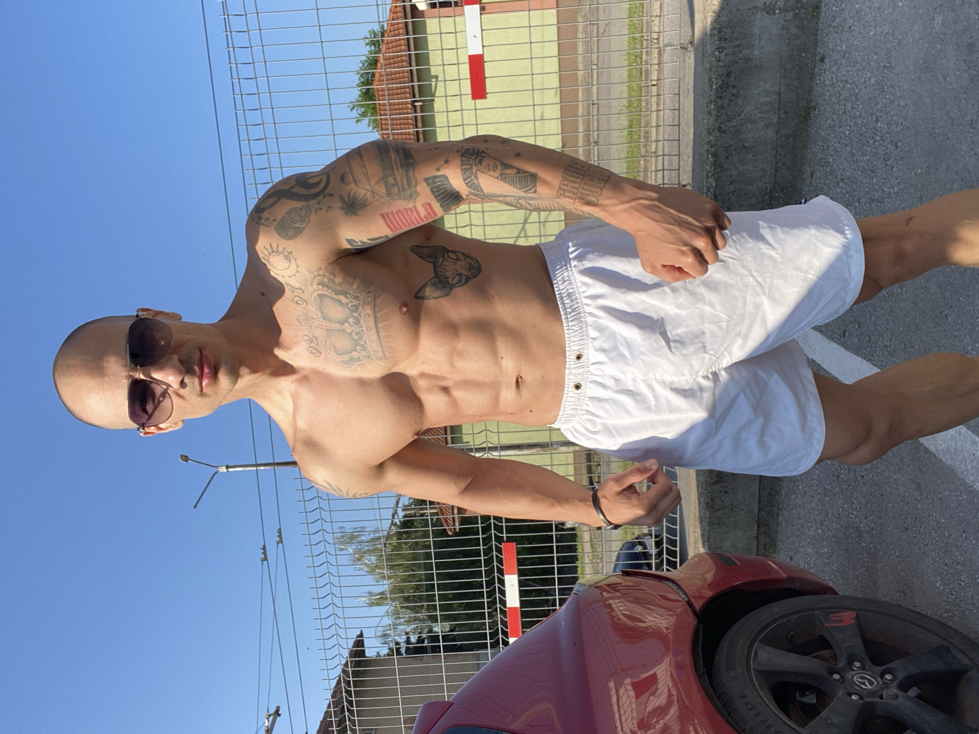 A muscular, bald man with numerous tattoos on his arms and torso poses shirtless in bright daylight. He wears dark sunglasses and white drawstring shorts, with a black wristband on his left arm. He stands directly facing the camera, with a serious expression.
Notable tattoos include the word "WORLD" and an Egyptian-style figure on his right arm, and a Sphynx cat on his left pectoral. The word "HATED" is also faintly visible on his left arm, along with a sun tattoo on his right shoulder.
The setting is outdoors in Pleven, Bulgaria, under a clear blue sky. A wire mesh fence runs horizontally behind the man. Beyond the fence, a house with a red-tiled roof and a light green wall is visible, along with trees and a red and white striped barrier pole. In the lower right foreground, the red fender and black wheel of a car are visible, with the number "3" highlighted in red on the wheel. The man is standing on a paved surface, likely asphalt. The harsh shadows suggest it is mid-day.