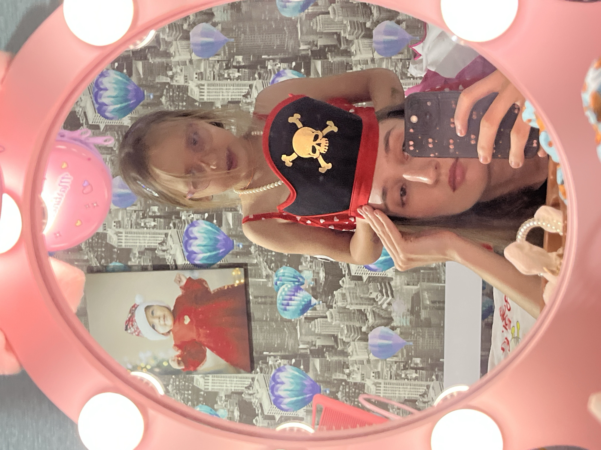 In a close-up selfie reflected in a pink-rimmed vanity mirror, a young girl with blonde hair and glasses wears a pirate hat with a skull and crossbones emblem. She is positioned behind a woman whose face is partially visible in the foreground. The background behind the subjects displays a cityscape wallpaper featuring hot air balloons. The woman is holding a smartphone, taking the photograph.