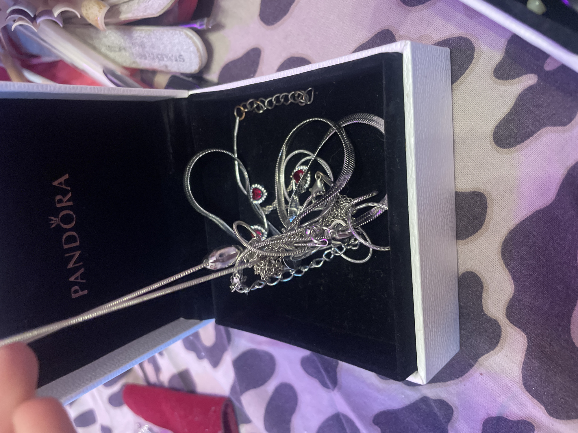 A silver Pandora necklace with two red heart-shaped charms is resting inside an open white and black Pandora jewelry box. The box is placed on a surface with a cow print pattern. The background is blurred, but suggests the presence of other items. The image does not contain information about the city of Ladozhskoe Ozero, Russia.