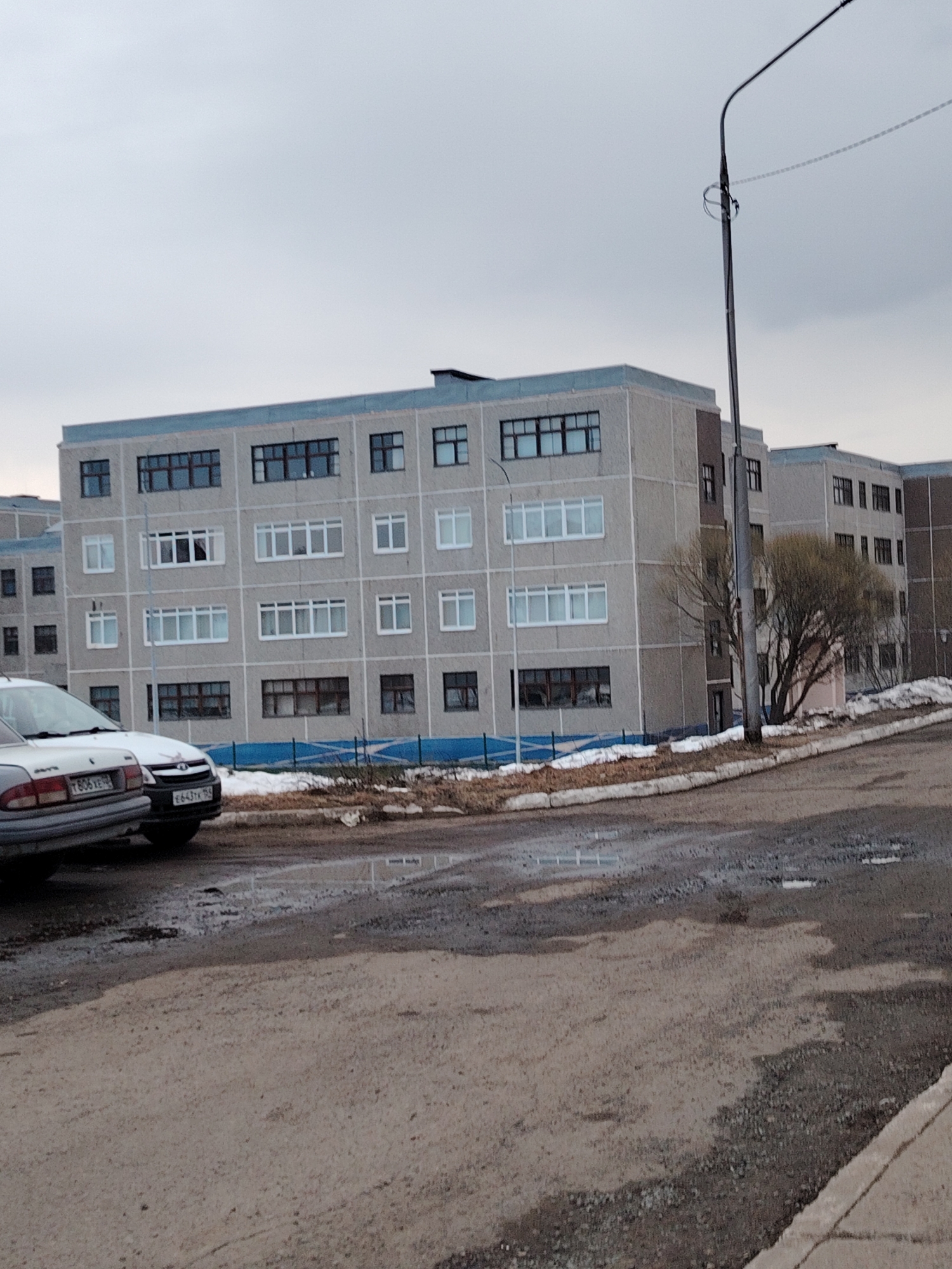 The scene depicts a residential area in Markovskii, Russia, characterized by grey, multi-story apartment buildings. The buildings have a repetitive facade of windows and lack distinctive architectural features. The foreground features a cracked asphalt road with visible puddles and patches of gravel. Two cars are parked near the curb. A bare tree stands adjacent to the building, and a utility pole extends into the sky. The overall atmosphere is overcast and drab. The presence of residual snow indicates a cold climate. No people are visible. The scene conveys a sense of urban decay and bleakness.
