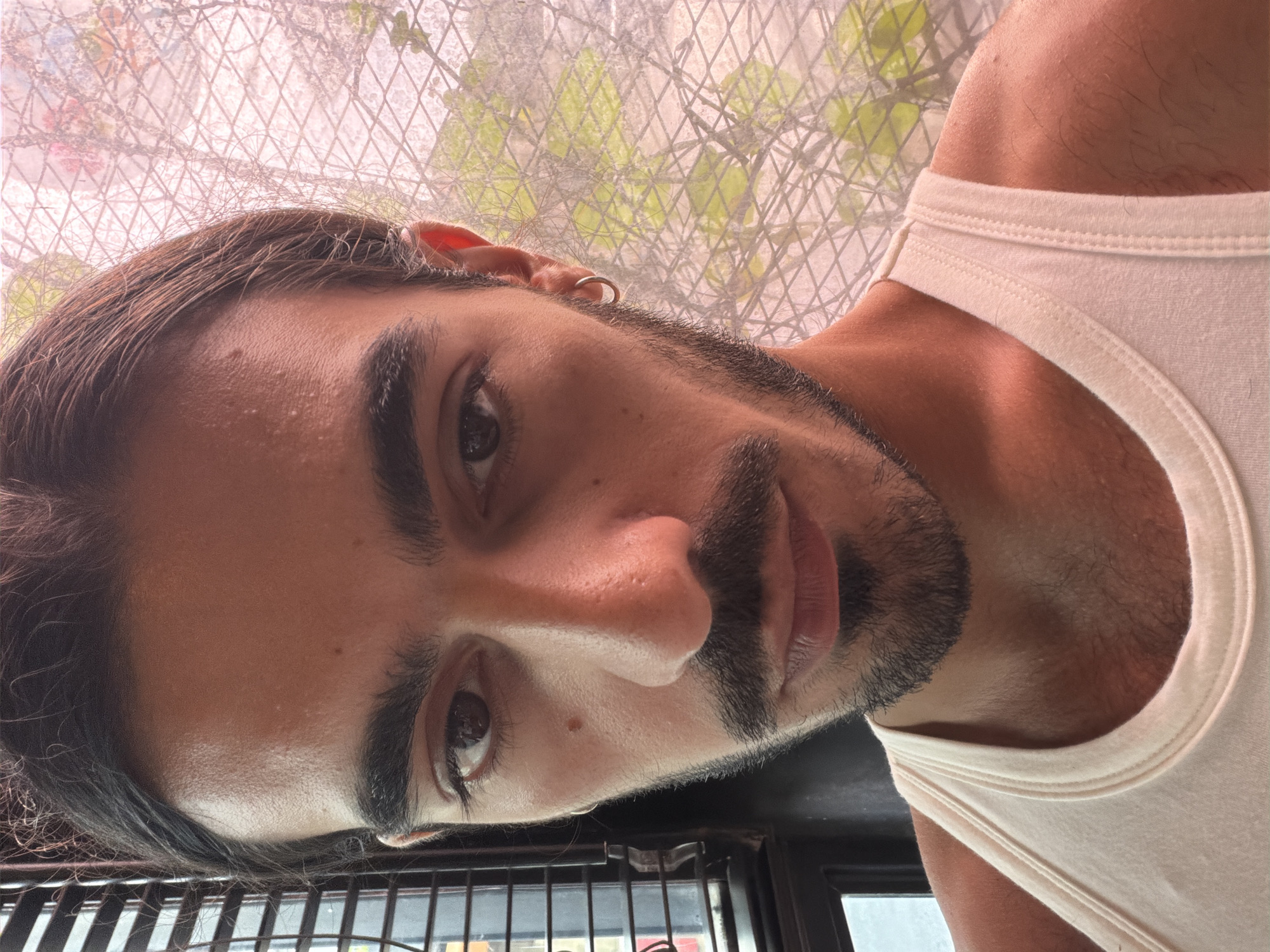 A close-up, upward-angled shot shows a man with dark hair pulled back from his face. He has dark eyes, a prominent nose, and a beard. He is wearing a white tank top. The background shows a window with a metal screen and blurred foliage. The location, North Kuta, Indonesia, is mentioned.