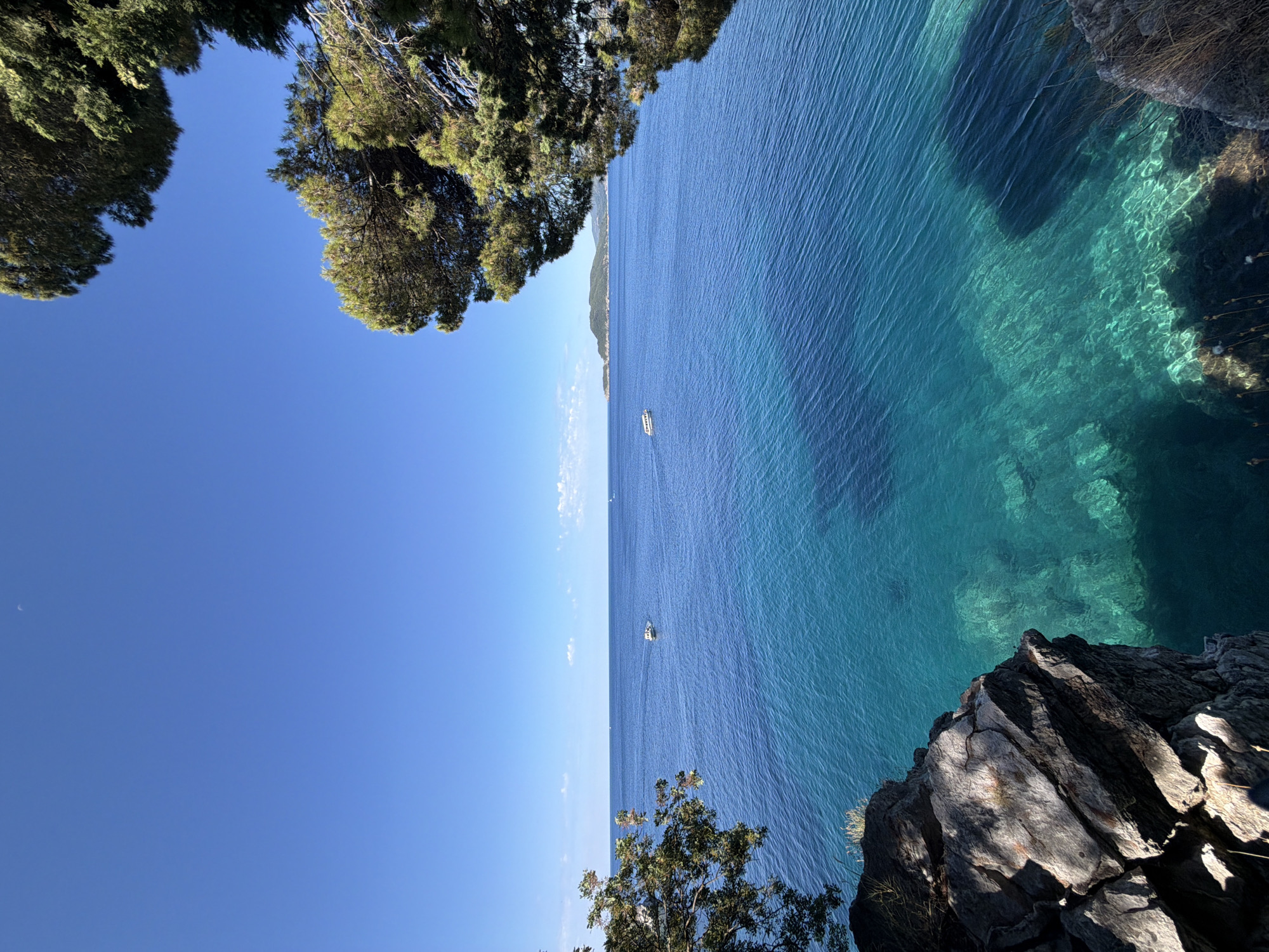 The image captures a stunning coastal view in Sveti Stefan, Montenegro. In the foreground, large, grey rocks are visible on the left, partially obscured by lush green foliage. To the right, the vibrant, turquoise waters of the Adriatic Sea dominate the scene, revealing a clear view of the rocky seabed beneath the surface. The water transitions to a deeper blue further out towards the horizon. Several small white boats are dotted across the sea, and in the distance, a green, mountainous island can be seen under a bright, clear blue sky. The scene is framed by the shadows and branches of large evergreen trees in the upper right and left corners of the image.