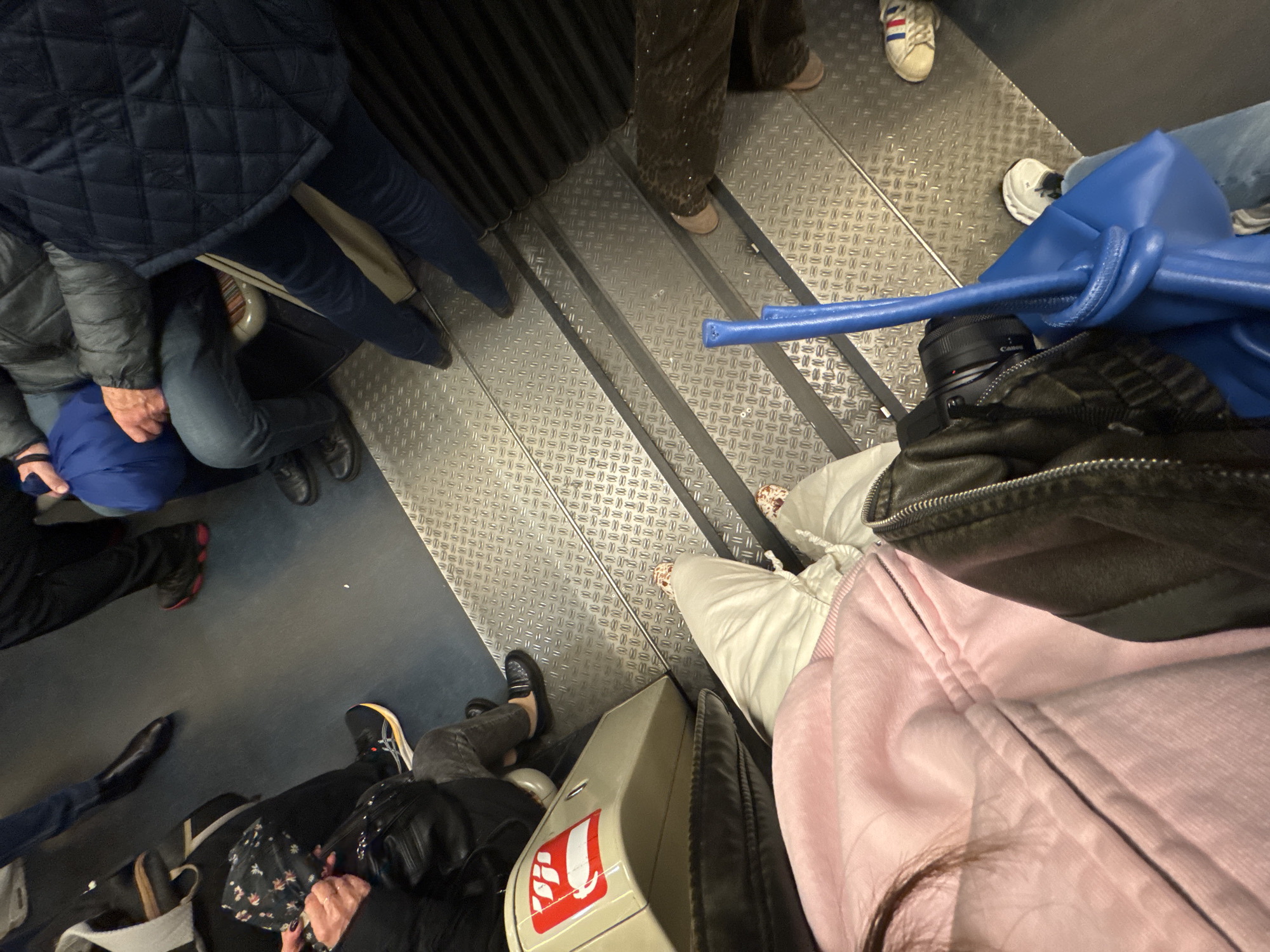 The image shows an interior shot of what appears to be a carriage on a train or metro. The floor is metal with a textured surface, and some areas show blue flooring. Multiple people are present, their feet and lower legs visible. Some individuals are standing on the metal floor, while others stand on the blue flooring. A blue bag and a camera lens are visible. Baggage is positioned around the area. The scene depicts a crowded public transportation environment. No specific landmarks confirming the location in Paris, France, are seen.