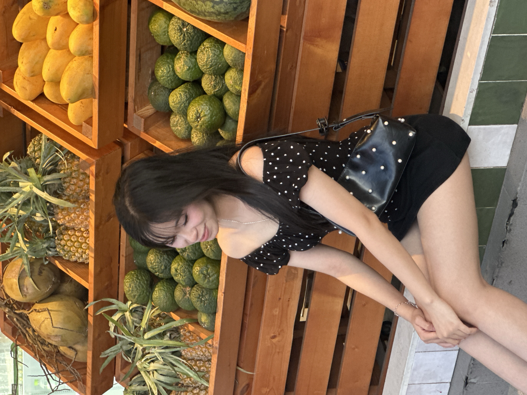 This media file captures a young woman with long, dark hair bent over a wooden stall displaying a variety of fresh fruits. The woman is wearing a black polka-dot top and a black skirt. She has a small black handbag with pearl accents slung over her shoulder. Her left arm is extended as she appears to be reaching for or examining the fruit, and her right hand is resting on her knee. A delicate silver bracelet adorns her left wrist.
The stall is divided into sections, each filled with different produce. One section showcases a vibrant display of ripe mangoes. Another section is packed with green, round fruits, likely avocados, stacked neatly. Below these, pineapples with their spiky green tops are visible, along with a few coconuts. The wooden structure of the stall suggests an outdoor market or a roadside fruit stand.
The lighting indicates it is likely late afternoon or early evening, with warm sunlight casting long shadows and highlighting the textures of the fruits and the wood. The background includes a glimpse of a green and white tiled wall, and a concrete surface, suggesting an urban setting. The overall mood is relaxed and casual, with the woman appearing to be enjoying her time at the market. There is no visible text in the image. The location context provided is Đà Nẵng, Vietnam.