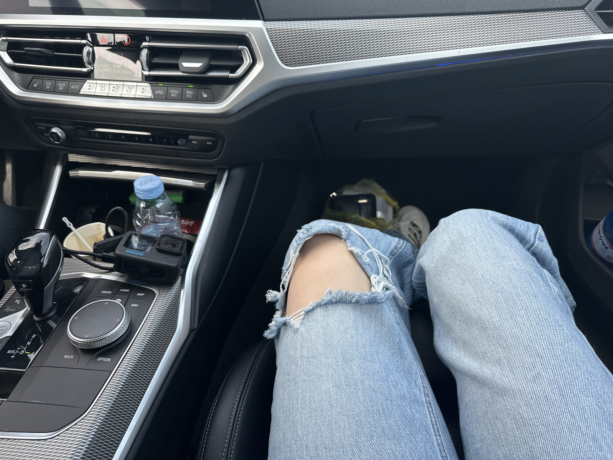 The media file captures the interior of a modern car, likely a BMW, from a first-person perspective, focusing on the passenger side footwell and parts of the dashboard and center console. A person's lower body is visible, clad in light blue, distressed jeans with a large tear at the knee, and light-colored sneakers, suggesting they are seated in the passenger seat.
The scene takes place inside the car, showing details consistent with a contemporary vehicle. The dashboard features air vents, climate control buttons, and a hazard light button. A blue ambient light strip runs along the textured silver trim above the glove compartment. The center console displays the gear shifter, an infotainment control knob, and various buttons.
Notable objects include a black GoPro camera (likely a Hero model) connected by a cable, a plastic water bottle with a blue cap, and a white cup, all placed in the console area near the gear shifter. The lighting appears bright, indicating it is daytime. The location context provided is Madrets, Bulgaria.
Visible text includes numerous labels on the car's controls: "MODE", "P", "OFF", "START ENGINE", "M/S D", "REAR", "AUTO", "MENU A/C", "MAX A/C", "MEDIA", "HOME", "MAP", "NAV", "BACK", and "OPTION". The GoPro logo is also identifiable on the camera. A partially visible label on the water bottle reads "VIN".