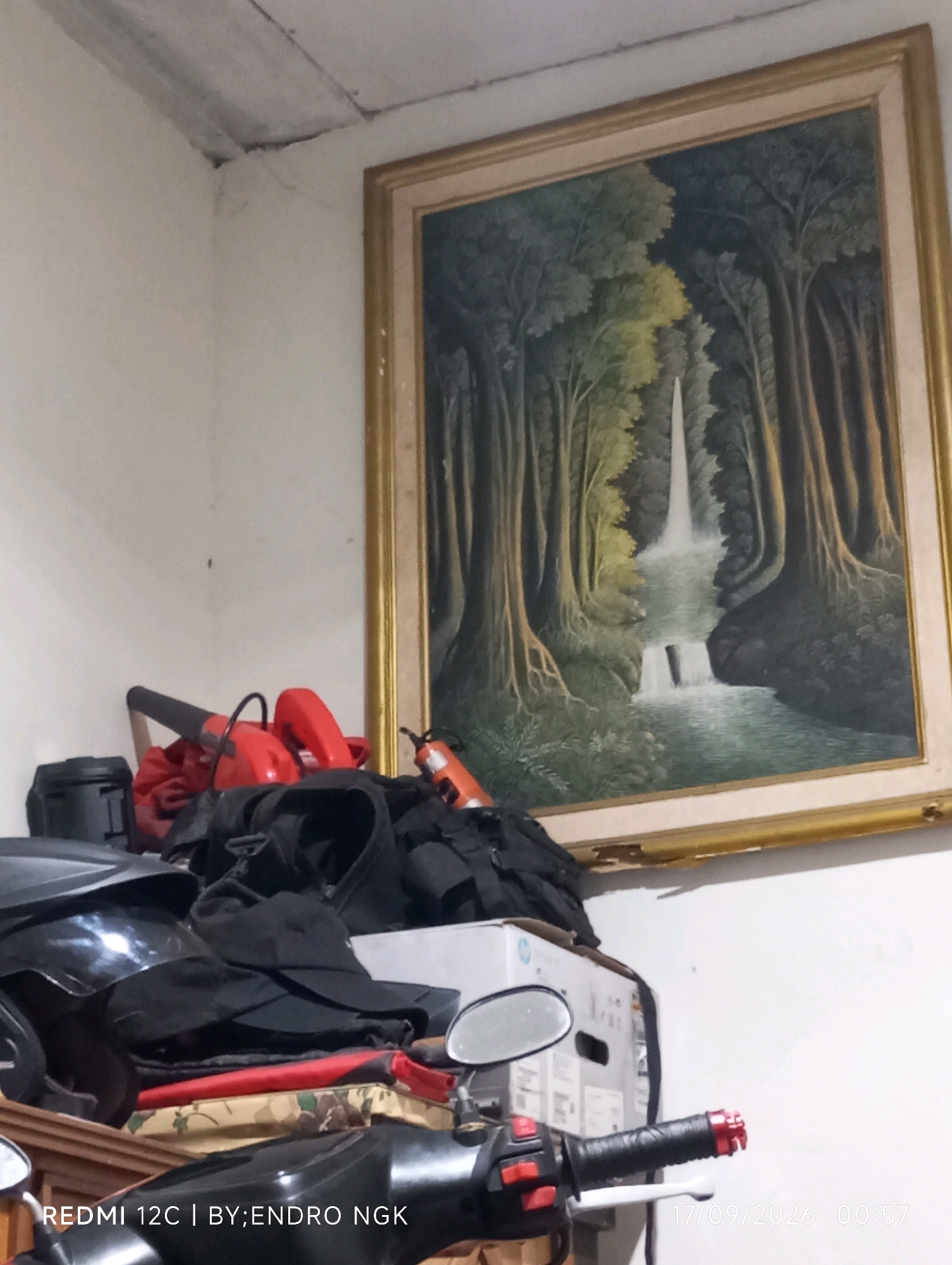 A painting of a waterfall in a forest is hung on a wall in a room. Below the painting are various objects, including a black helmet, a red leaf blower, and piles of black clothing. In the foreground, part of a motorcycle is visible, with handlebars and mirrors. The room appears to be cluttered, and the lighting is dim. The lower left corner of the image displays text that reads "REDMI 12C | BY:ENDRO NGK" and the date "17/09/2026" followed by the time "00:57". This image appears to have been taken in Ringinanom, Indonesia.