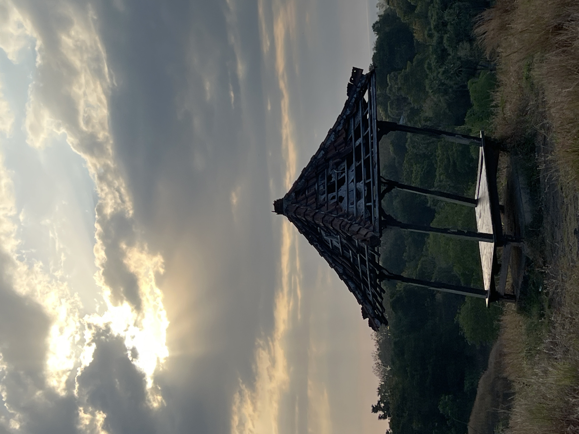 The image shows a dilapidated open-air pavilion on a grassy hilltop overlooking a dense forest. The sky above is filled with dramatic, textured clouds, with bright sunlight filtering through in rays, suggesting either sunrise or sunset. The pavilion itself is constructed of dark wood and has a tiled roof that appears to be partially collapsed or missing tiles, revealing the underlying structure. A wooden platform is visible within the pavilion. The foreground is covered in dry, tall grass, indicating a natural, perhaps somewhat overgrown, environment. The background features a thick canopy of green trees that descends down a slope, leading to what appears to be a distant body of water or coastline. The overall scene evokes a sense of isolation and natural beauty, with the decaying pavilion adding a touch of melancholy or historical resonance to the landscape. The specific location in Kubutambahan, Indonesia, implies a tropical or subtropical setting, which is consistent with the lush greenery and the appearance of the sky. There are no people or animals visible in the image.