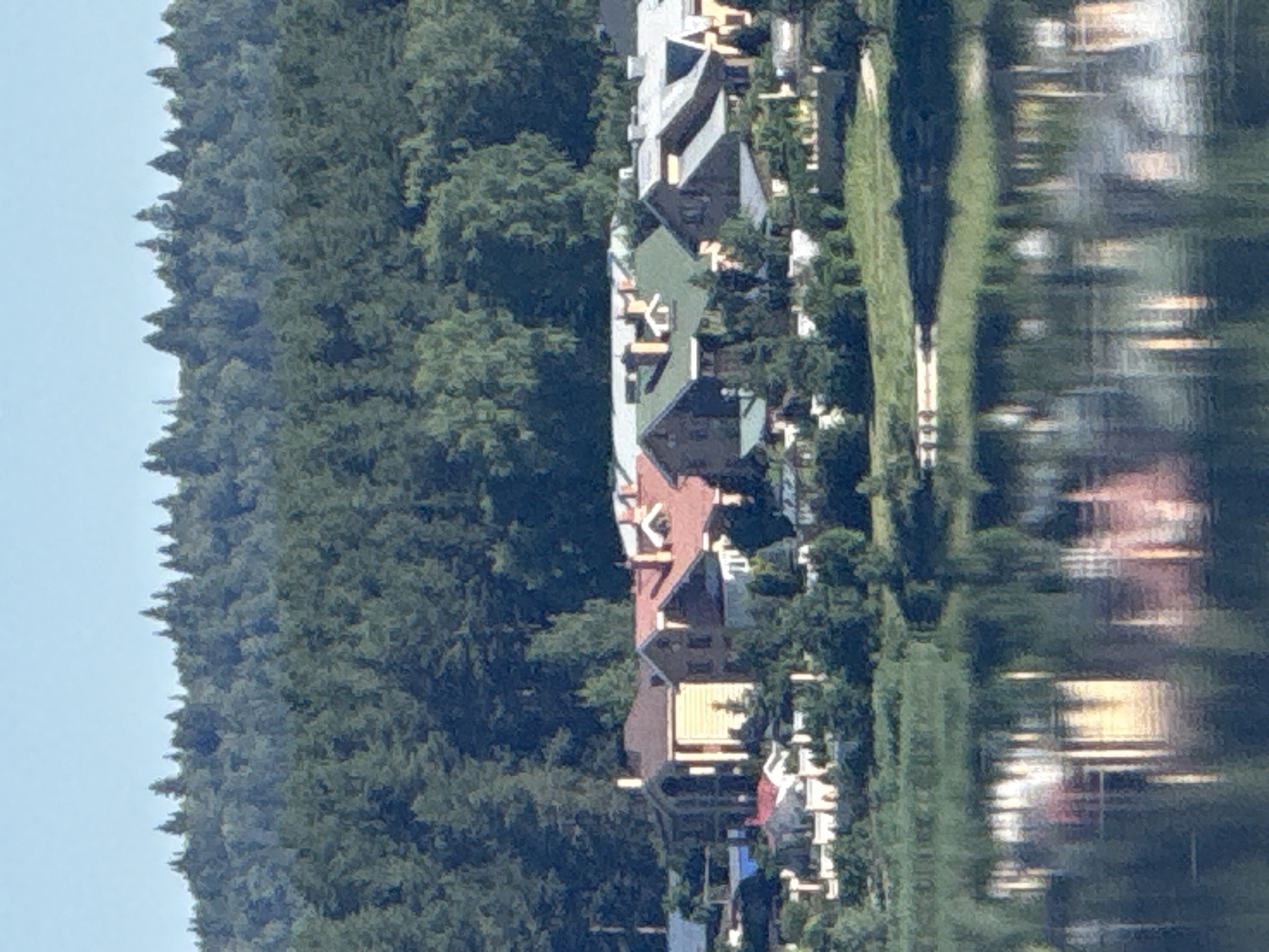 The image captures a panoramic view of a lakeside residential area with a dense forest backdrop, situated in Votkinsk, Russia. In the foreground, a tranquil body of water reflects the houses and the sky. The midground is dominated by a cluster of upscale houses, characterized by diverse architectural styles, red and green tiled roofs, and light-colored walls. These homes are nestled amongst lush greenery and trees. The background features a vast, undulating forest of dark green coniferous trees, stretching across the horizon under a clear blue sky. The scene depicts a serene, affluent, and natural setting, typical of a suburban or semi-rural environment.