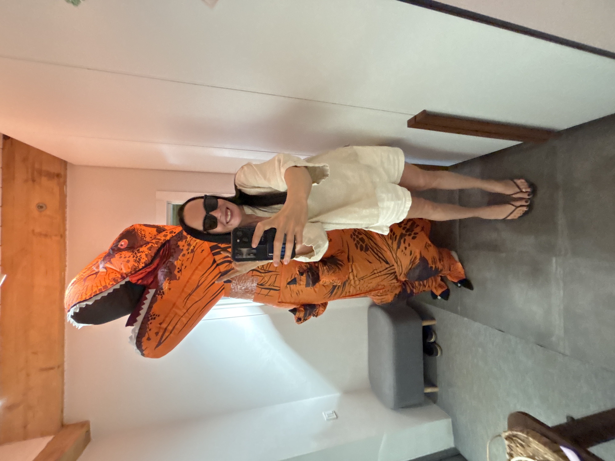A woman wearing a white dress and sunglasses is posing for a selfie in an orange inflatable dinosaur costume. She is holding a smartphone to take the photo. The location appears to be indoors, possibly a home interior. The background includes a light-colored wall, a wooden frame, and a gray ottoman with shoes underneath. The floor is a light gray color. The scene does not immediately suggest any specific location within the city of Chaikovsky, Russia.