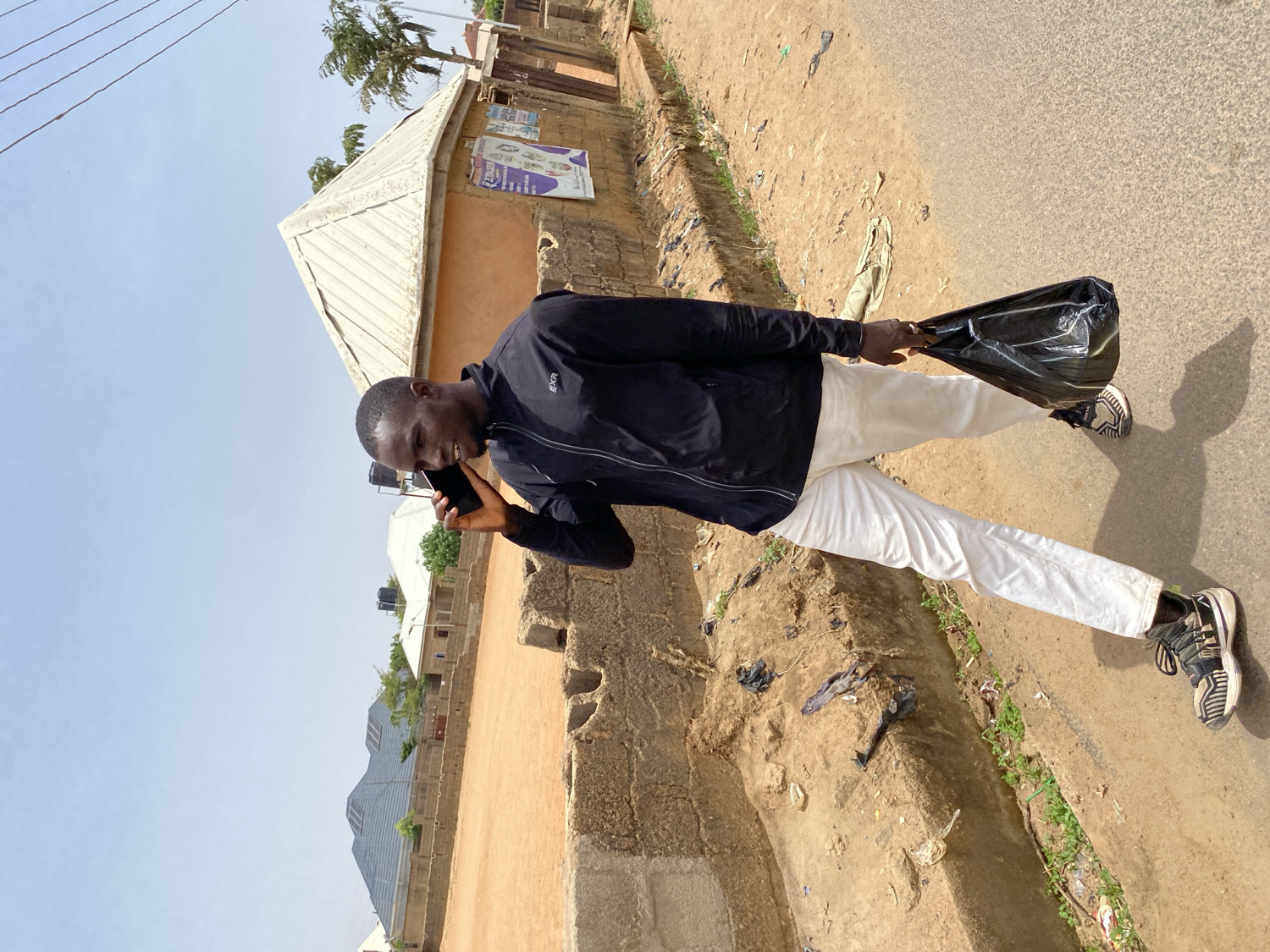 A young Black man is depicted standing on a paved road under a clear, bright sky. He is wearing a black long-sleeved top, possibly a jacket, with a faint white logo (appearing as "EXR" or similar) on the left chest, white trousers, and black and white sneakers. In his left hand, he holds a black plastic bag, while his right hand holds a black smartphone to his ear, suggesting he is talking on the phone. A slight smile is visible on his face.
The scene is set during bright daylight, indicated by distinct shadows and a clear blue sky, implying midday or early afternoon. To his right, the paved road transitions into a dry, unpaved area of reddish-brown earth, which contains sparse green weeds and scattered litter, including discarded plastic. In the background, there are low-rise buildings with corrugated metal roofs. One building's wall features posters or advertisements, the text of which is illegible. Further in the distance, more structures and a large water tank are visible under the expansive sky. The environment appears dry and dusty, characteristic of a semi-urban landscape in a sunny climate, aligning with the given context of Jos, Nigeria.