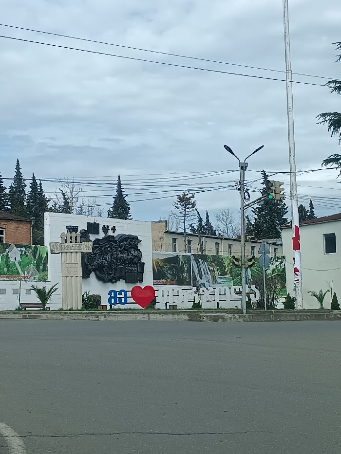 The image depicts a street scene in the city of Terjola, Georgia, under an overcast sky. In the foreground, an asphalt road with a partial white lane marking is visible. The central feature is a white, rectangular monument wall adorned with various elements.
On this wall, a prominent black bas-relief sculpture depicting multiple figures is centrally positioned. To the left of the bas-relief, a tall, fluted white pillar-like structure with a decorative top and some illegible text is present. Below the bas-relief, a large red heart symbol is flanked by blue Georgian characters. Further to the right of the heart, more white Georgian characters are visible along the base of the wall.
Two large banners or murals are affixed to the wall: one on the right featuring a cascading waterfall in a lush landscape, and another on the far left displaying a green, terraced landscape. A brown wooden bench is situated at the base of the wall on the far left, next to a small, palm-like plant. Several small green shrubs are planted along the base of the monument.
To the right of the monument, a tall utility pole supports a lamppost with two lights and a traffic light showing a green signal. Numerous black power lines crisscross the upper portion of the frame. A slender, tall white pole stands to the right of the utility pole, adjacent to a white building partially obscured by a vertical red and white striped element, possibly a flag or banner. A blue circular road sign with a white arrow is visible behind the lamppost.
In the background, behind the monument, multiple trees are visible, including tall, dark conifers and bare deciduous trees. Several buildings are present, including a light-colored multi-story structure with multiple windows directly behind the monument, and a brick building on the far left. No individuals or active interactions are discernible.