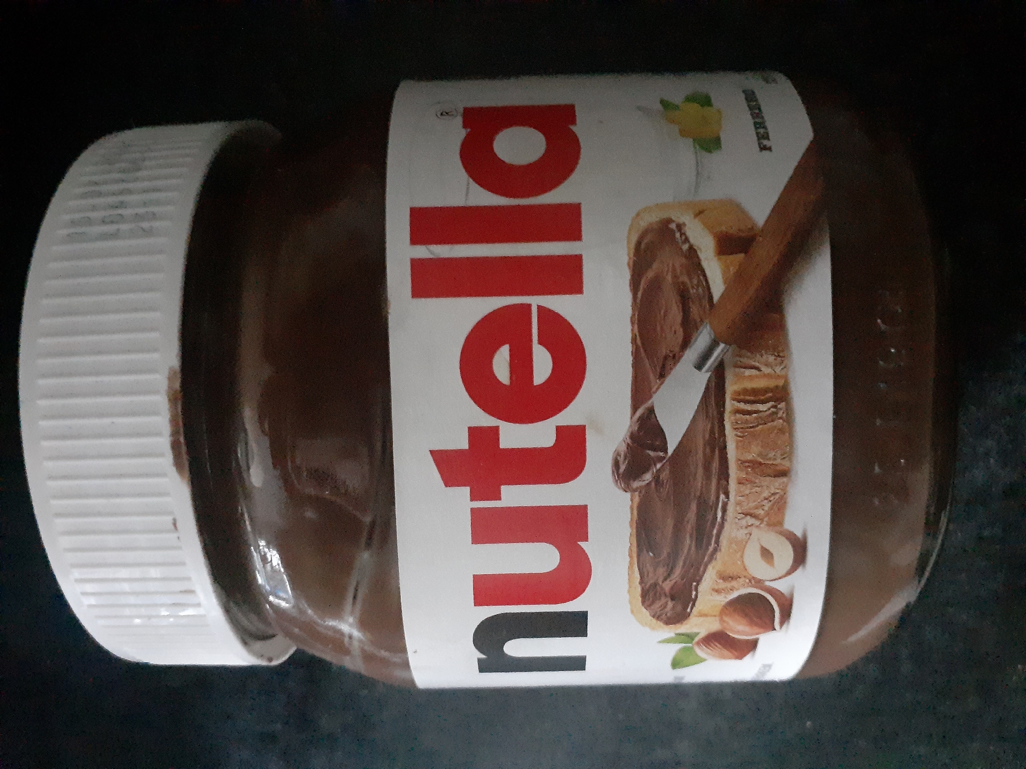 An overhead medium shot shows a jar of Nutella on a dark, textured surface. The jar is mostly filled with the chocolate hazelnut spread and has a white ridged lid. The label on the jar is white with red lettering that spells "nutella." Below the name, there is an image of a slice of bread spread with Nutella, with a knife and some hazelnuts. To the right of the image on the bread, the word "FERRERO" is visible in green lettering. The dark surface appears to be a countertop.