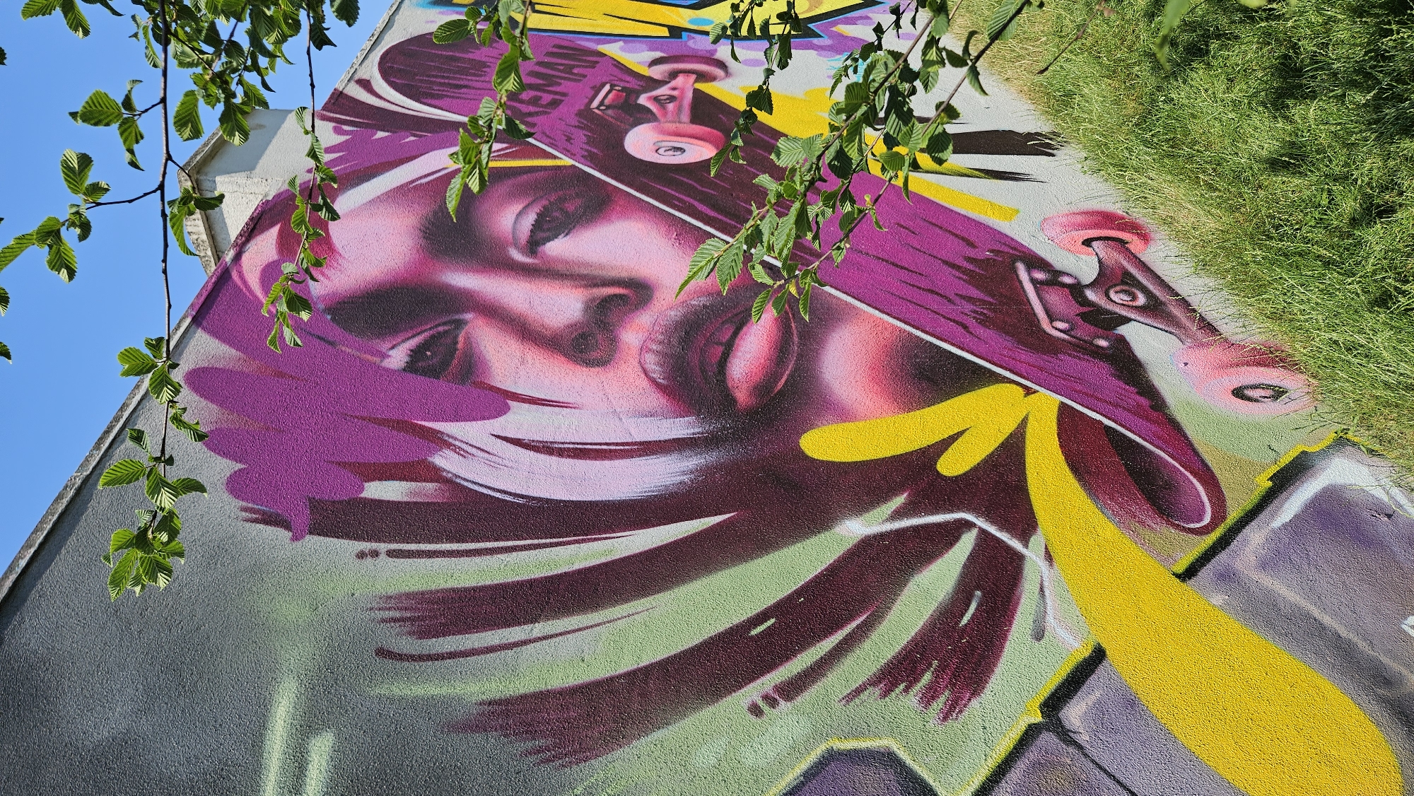 An outdoor mural is painted on a grey concrete wall. The mural depicts a close-up of a woman's face with striking purple and pink tones, her eyes closed or looking down. Her lips are full and painted in a deep red. A purple skateboard is positioned diagonally across the lower right portion of the mural, obscuring part of the woman's face. The skateboard has pink wheels and the word "TREKKEMAN" is visible in white lettering on its surface. The background of the mural features abstract shapes and streaks of color, including green, yellow, and purple. In the foreground, green leaves and branches from a tree hang over the top right and right sides of the mural, partially obscuring the view. Lush green grass grows at the base of the wall. The sky above is clear and blue. The mural is located on a wall along a street or pathway in Prešov, Slovakia, with a grassy slope adjacent to the wall.