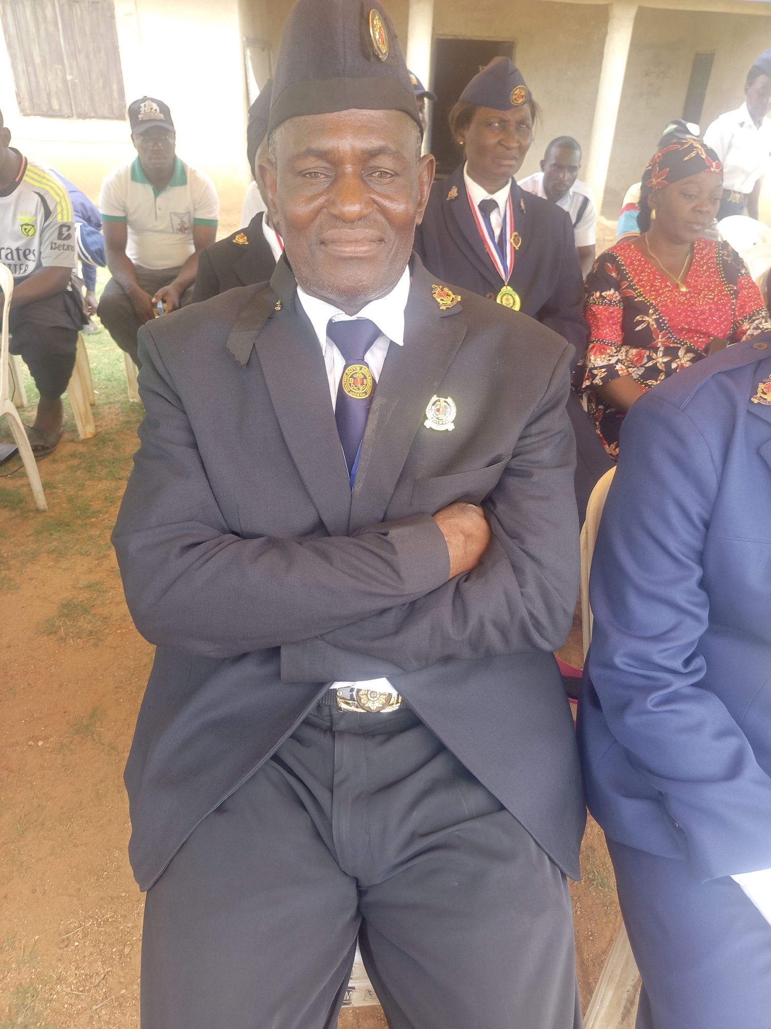 A man sits in Akwanga, Nigeria, wearing a dark suit and matching cap adorned with a logo. He has his arms crossed and a smile on his face. Behind him are other people seated, including a woman in a similar uniform with a medal, and another woman in a red and black patterned dress. The background is a dusty outdoor area with a building visible in the distance.