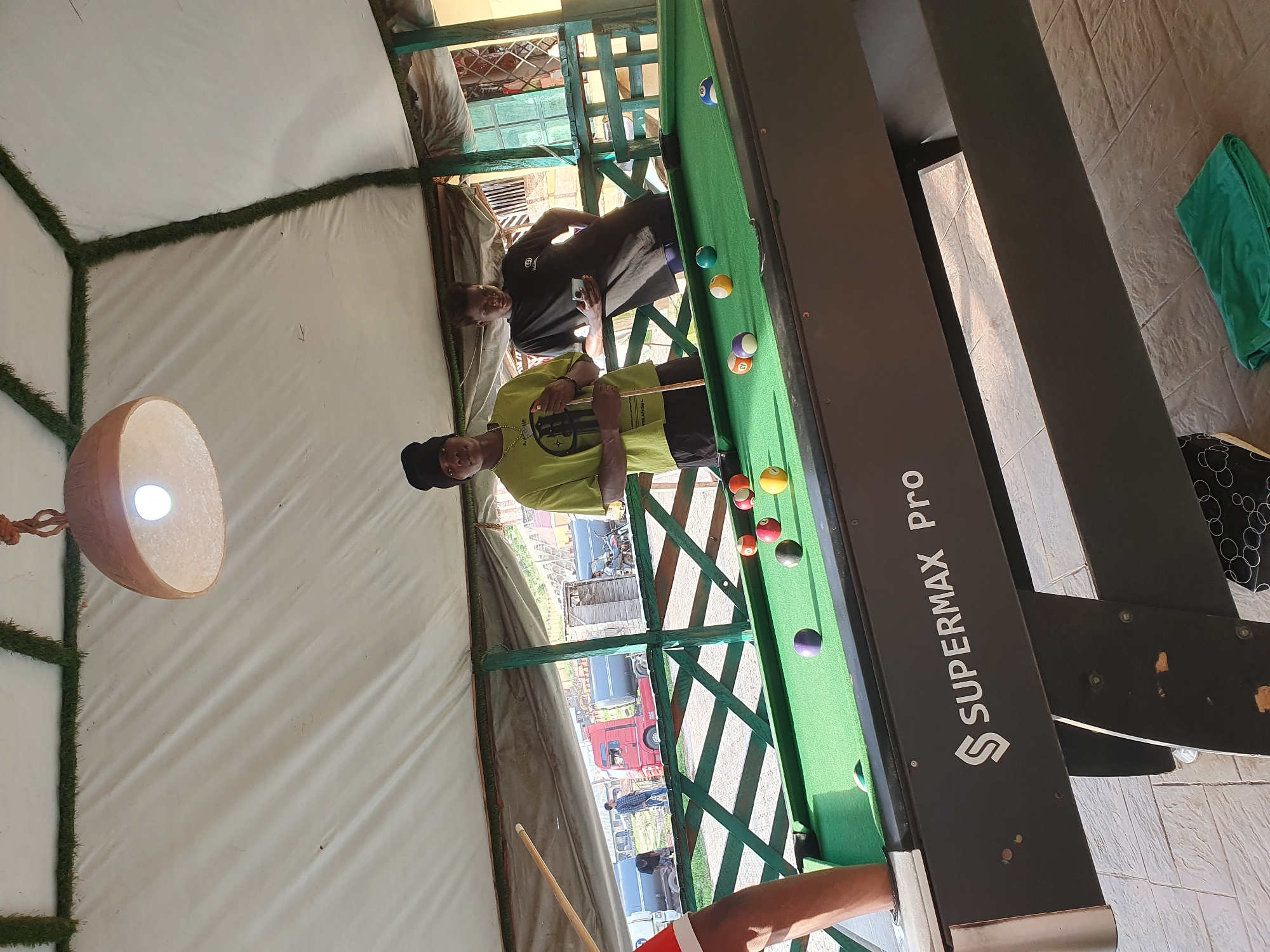 In Lokoja, Nigeria, two young men are playing a game of pool on a green felt table under a white canvas roof supported by green wooden beams. One man, wearing a black beanie and a light green t-shirt, stands to the left, holding a pool cue, and looking towards the camera. The other man, in a black t-shirt, stands to his right, looking down at his phone. Several billiard balls are scattered across the table, with one purple ball prominently displayed in the foreground. The pool table is labeled "SUPERMAX Pro" on its side. In the background, through a green lattice fence, a red truck and other vehicles are visible on a road, suggesting an outdoor, possibly commercial or recreational area. The ground beneath the table is tiled, and a green towel lies on the floor in the lower right corner. A light fixture hangs from the ceiling, illuminating the scene.