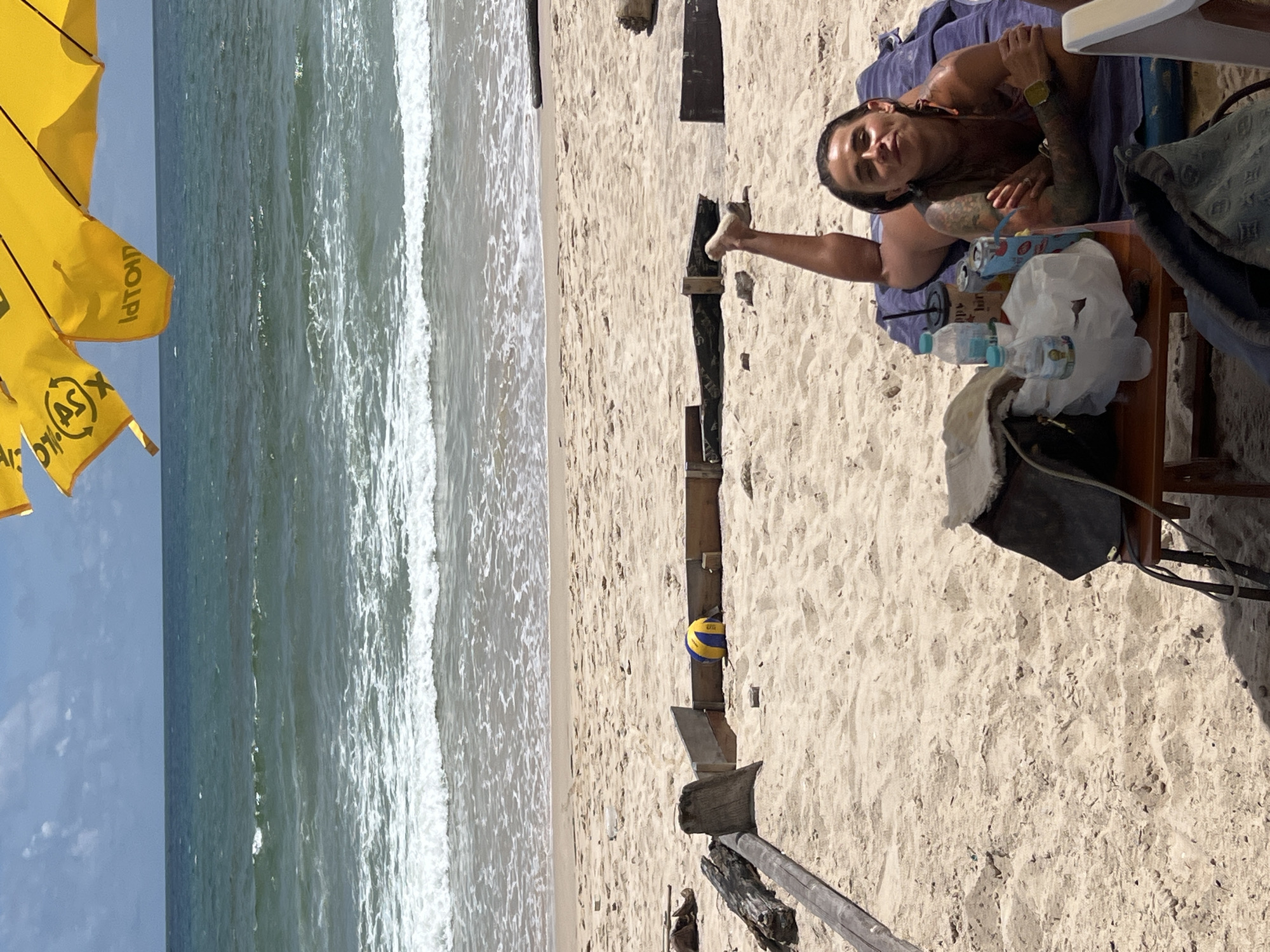 A woman is lying on a blue towel on a sandy beach, facing the camera with her legs extended. She has tattoos on her arms and is wearing a bikini top. In front of her is a small wooden table with drinks and bags. A yellow umbrella is visible at the top of the frame. The ocean and waves are in the background. The scene is set on a sunny day.