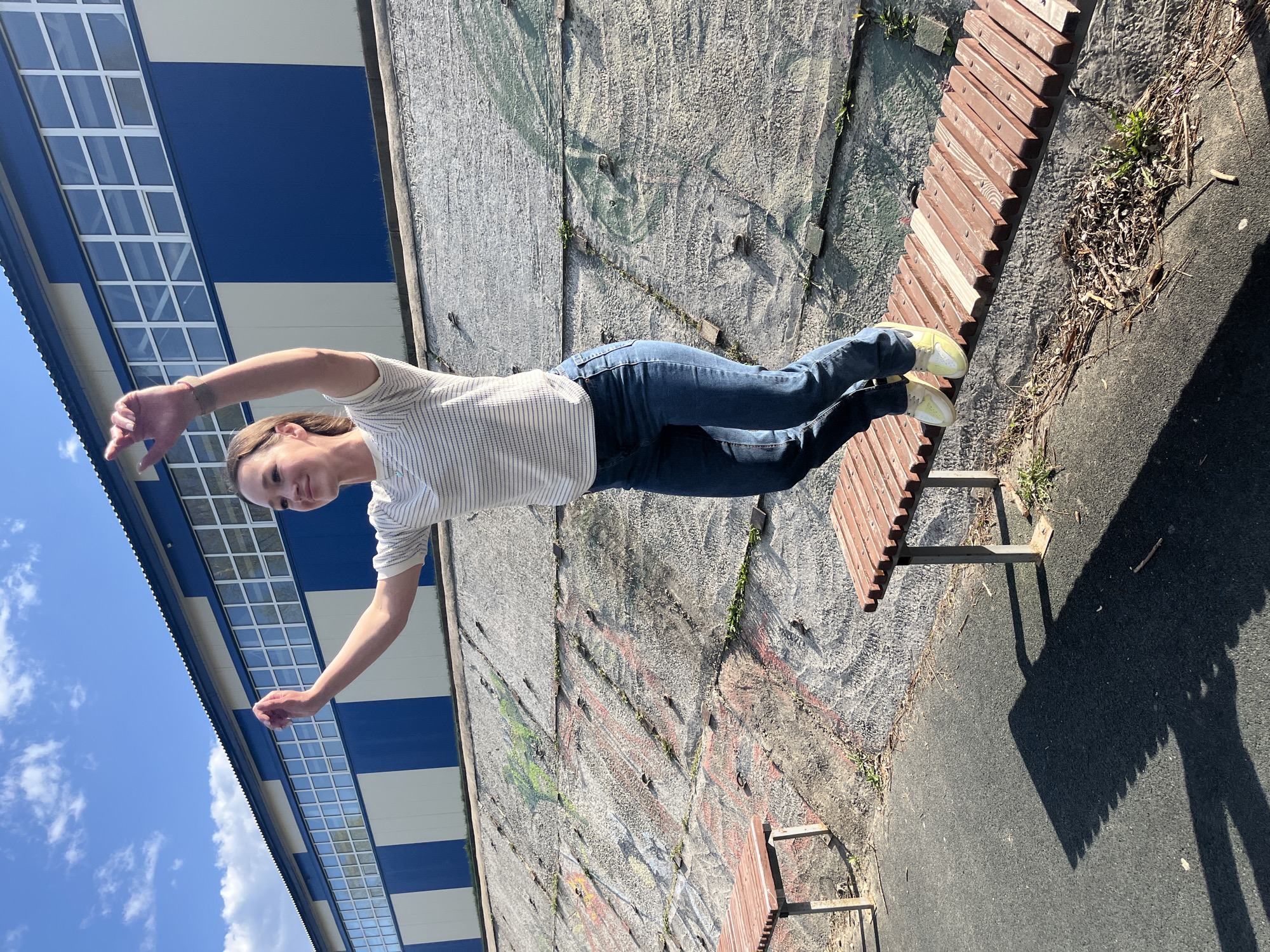 A woman is posing on a bench in an outdoor setting. She is wearing a striped shirt, blue jeans, and yellow shoes. Her arms are outstretched. A building with blue and white panels is visible in the background, alongside a graffiti-covered wall. The location appears to be a park or public space in an urban area. The scene is in daylight. The photo does not provide information that it is in Ulyanovsk, Russia.