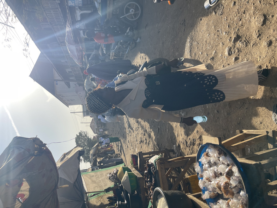 A woman wearing a light colored skirt, dark patterned top, and cream-colored jacket stands on a dirt road, holding a water bottle. She is situated in what appears to be a crowded outdoor market or street. Vendors' stalls are visible, one with items packaged in clear plastic. Motorbikes and other vehicles are parked alongside the road, with buildings and more activity in the background. The scene is bathed in bright sunlight.