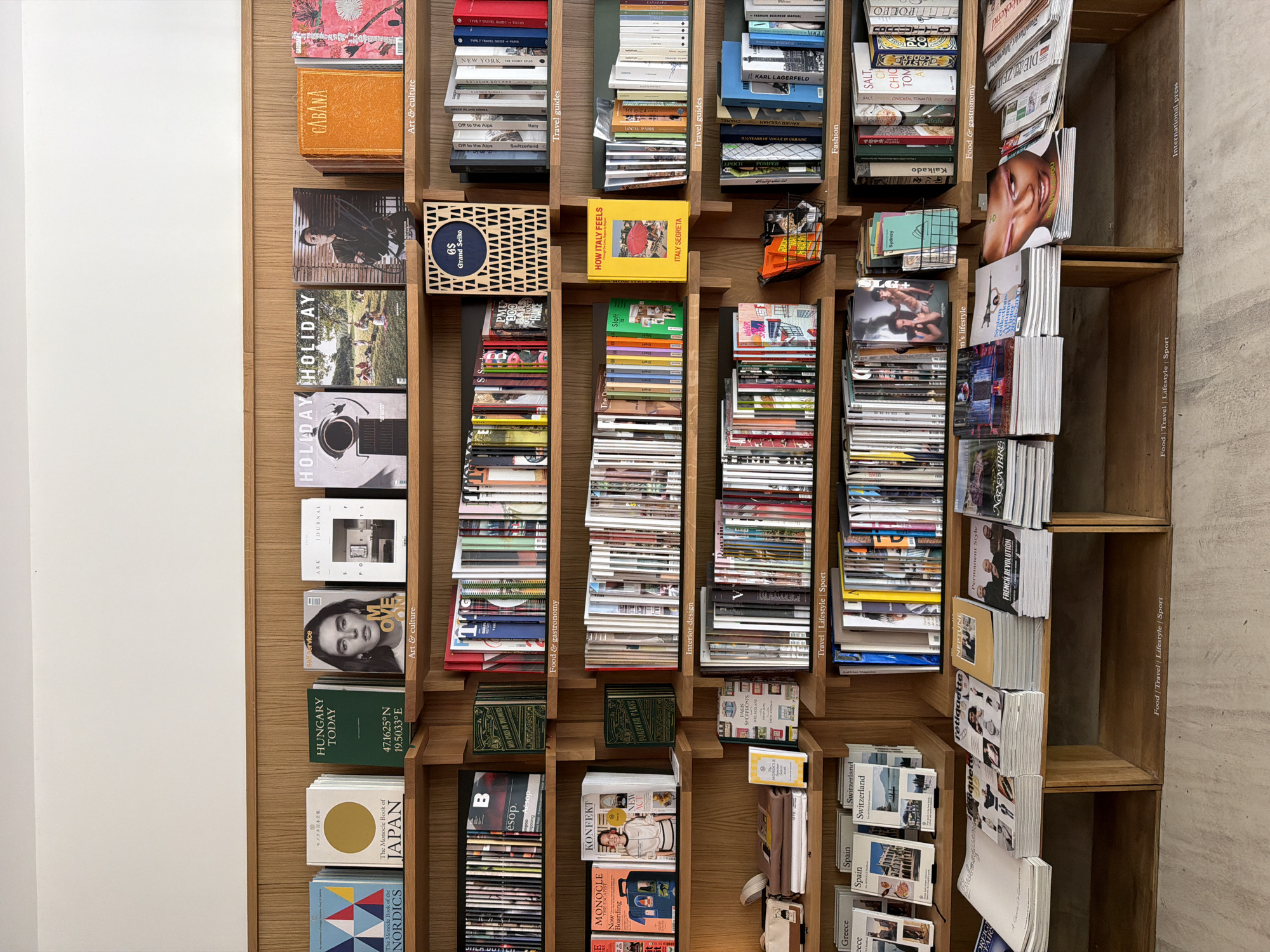 The image shows a wooden bookshelf filled with a variety of books and magazines. The publications cover topics such as travel guides, food and gastronomy, interior design, fashion, lifestyle, and art & culture. Some prominent titles include "Cabana", "Holiday", "Hungary Today", "The Monocle Book of Japan", "How Italy Feels", "New York, The Secret Atlas", and "Karl Lagerfeld". The books are arranged vertically and horizontally within the shelves. Some shelves also contain additional items like small boxes and pouches. Text on the shelves denotes thematic sections such as "Art & Culture", "Travel Guides", "Men's Lifestyle", "Fashion", "Food & Gastronomy", "Interior Design", "Travel Lifestyle Sport", and "International Press". The bookshelf is positioned against a plain white wall, with the right side of the shelf against a beige wall. Based on the presence of publications such as “Japon-Paris De Toujours” and “Salon Paris”, this is likely in Paris, France.