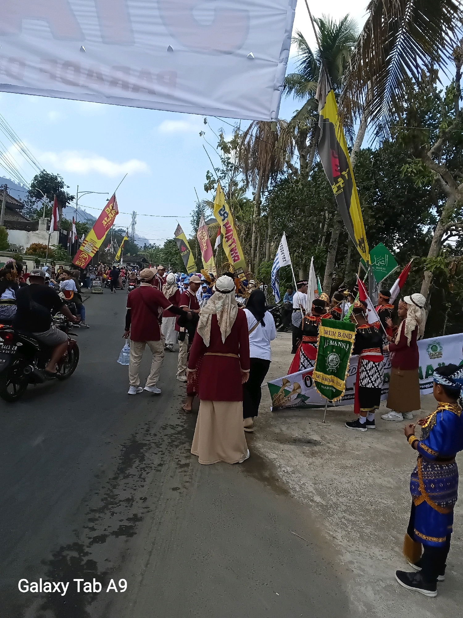 The image depicts a street scene in Bendo, Indonesia, likely during a parade or cultural event. A group of people are gathered, many dressed in traditional attire. Several individuals, including children and adults, are wearing ornate costumes featuring colorful fabrics and headdresses. Some are carrying flags and banners, some of which are emblazoned with text and symbols.
In the foreground, a young person in a bright blue and gold outfit stands facing away from the camera. To their left, other participants in red and tan uniforms are visible, some holding flags and a banner that reads "DRUM BAND AL MANNA". In the background, more people and a motorcycle are present, suggesting a public gathering. The street is paved and appears somewhat wet in places, with scattered debris. Palm trees and other vegetation line the street, and a large white banner stretches across the top of the frame. The sky is partly cloudy. The overall atmosphere suggests a festive and community-oriented event.