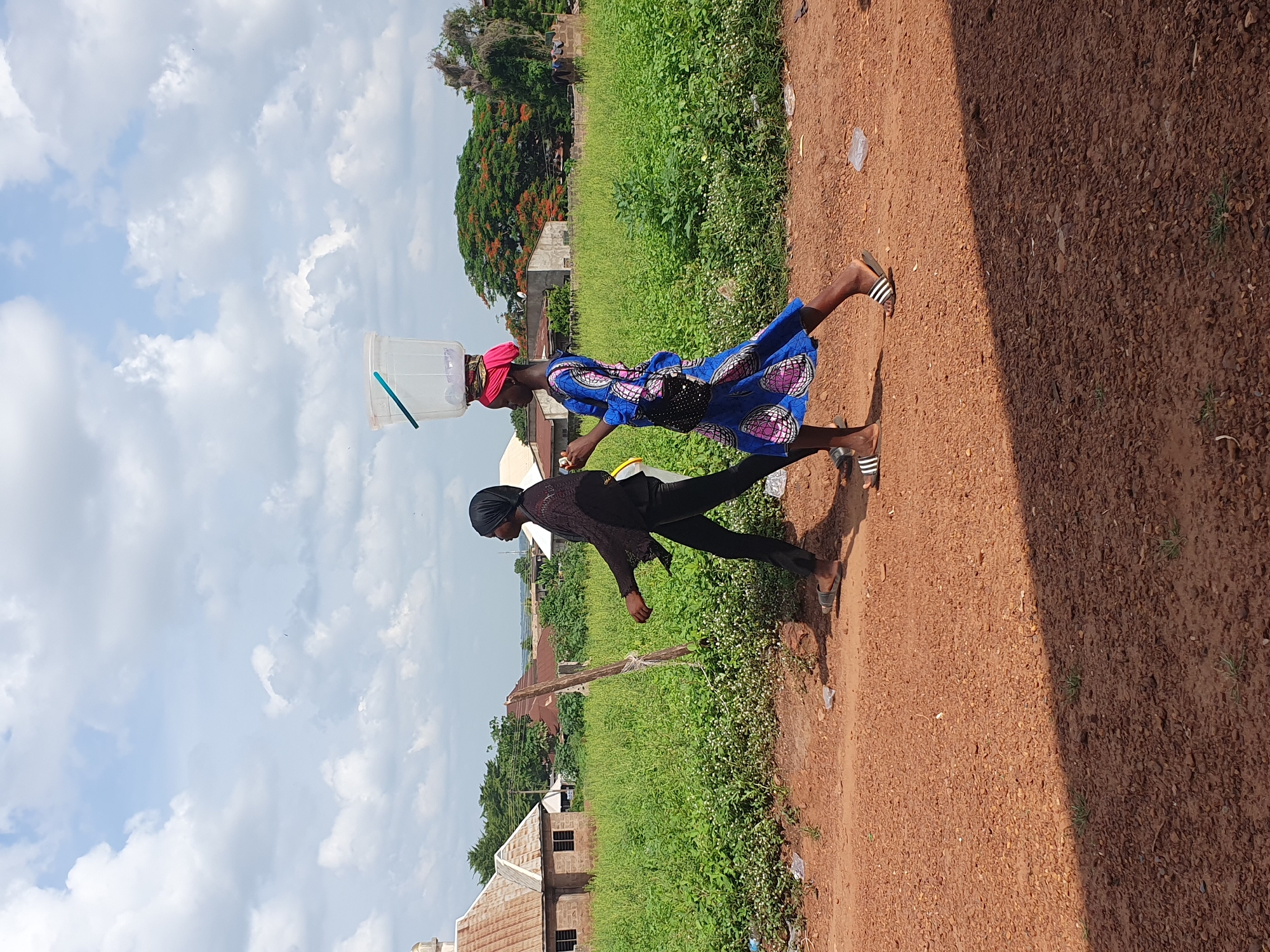 Two women walk along a reddish-brown dirt path in a rural or semi-urban setting in Idah, Nigeria. The woman in front carries a clear plastic container, possibly for water, balanced on her head. She wears a red headwrap, a vibrant blue dress with a pink and white pattern, and striped flip-flops. A small black patterned bag hangs from her waist. Behind her, the second woman walks in dark clothing, consisting of a black head covering, a dark long-sleeved top, and dark trousers, also wearing striped flip-flops.
To their left, beyond the path, is an expanse of lush green vegetation, including tall grasses and bushes. In the background, simple, single-story buildings with corrugated iron roofs are visible amongst trees. A prominent tree with reddish-orange blossoms (likely a Flamboyant tree) stands further back.
The sky is partly cloudy, with large white cumulus clouds against a bright blue sky, indicating bright daylight, likely midday or early afternoon. Strong sunlight casts distinct shadows on the dirt path. The scene depicts a routine activity, possibly fetching water or goods, in a warm, outdoor environment. There is no visible text in the image.