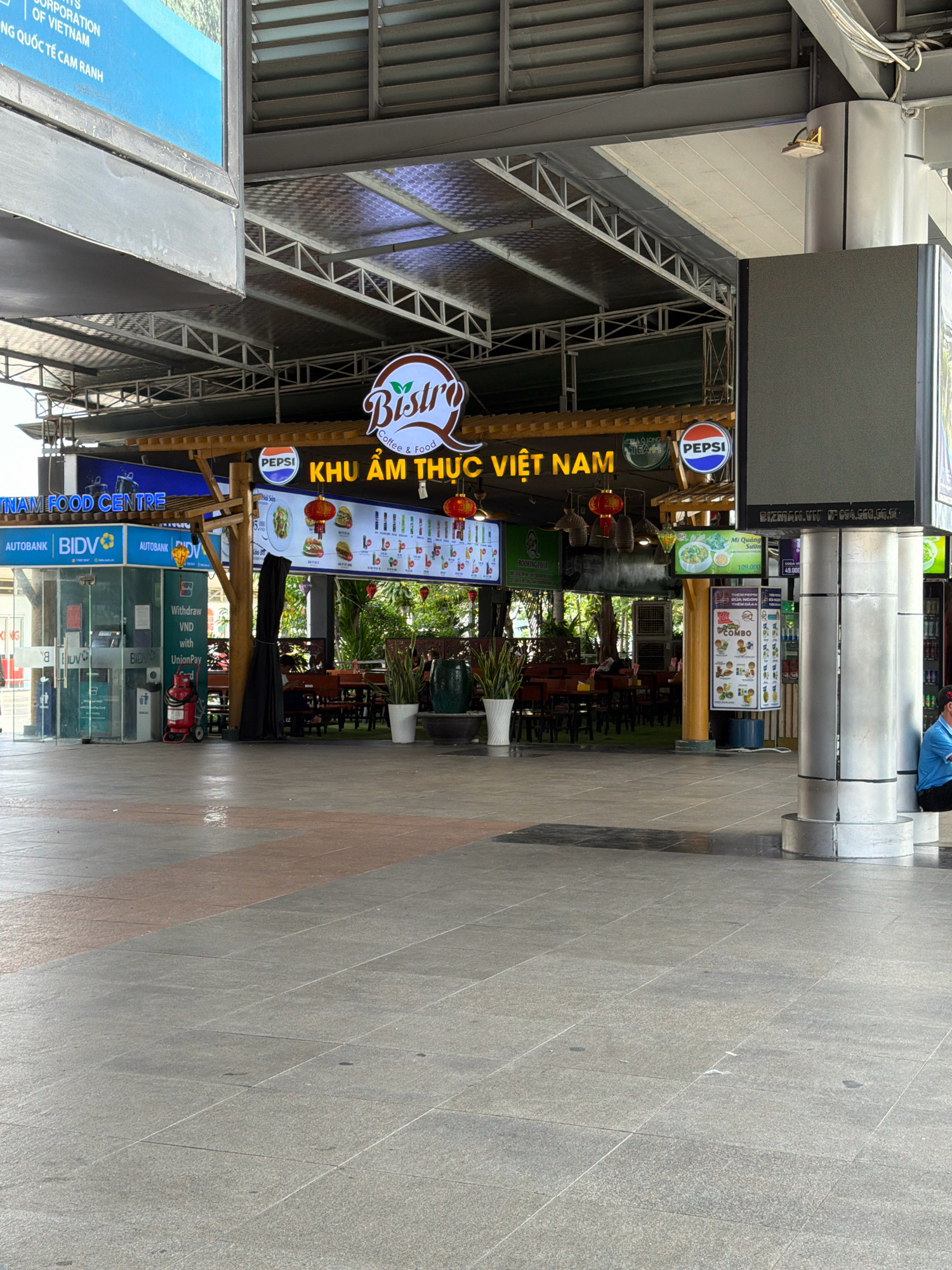 The image captures an outdoor commercial area in Bac Cam Ranh, Vietnam. In the foreground, a grey tiled plaza is visible, with some scattered debris and a wet patch reflecting the overhead structure. To the left, a glass-fronted building displays signage for "AUTOBANK" and "BIDV," indicating a banking facility. A red fire extinguisher is positioned outside this building.
The central area features a food court with a wooden canopy and a large illuminated sign that reads "KHU ẨM THỰC VIỆT NAM," translating to "Vietnamese Food Area." Above this sign, a "Bistro Coffee & Food" logo is prominent. Several smaller screens display menus with images of food items, including burgers and Vietnamese dishes. Red lanterns are suspended from the canopy, and Pepsi logos are visible on some signs. Behind the food court, tables and chairs are set up for dining, with some potted plants and greenery visible.
To the right of the food court, a large metallic cylindrical pillar supports an overhead structure. A large, dark, rectangular digital screen is mounted on a pole to the right of this pillar, displaying an advertisement. A person in a blue shirt is seated on the ground next to the pillar, facing away from the camera. The overall environment suggests a bustling commercial zone, likely a transportation hub or a shopping center, given the presence of banking services and a food court catering to travelers or shoppers. The lighting suggests daytime.