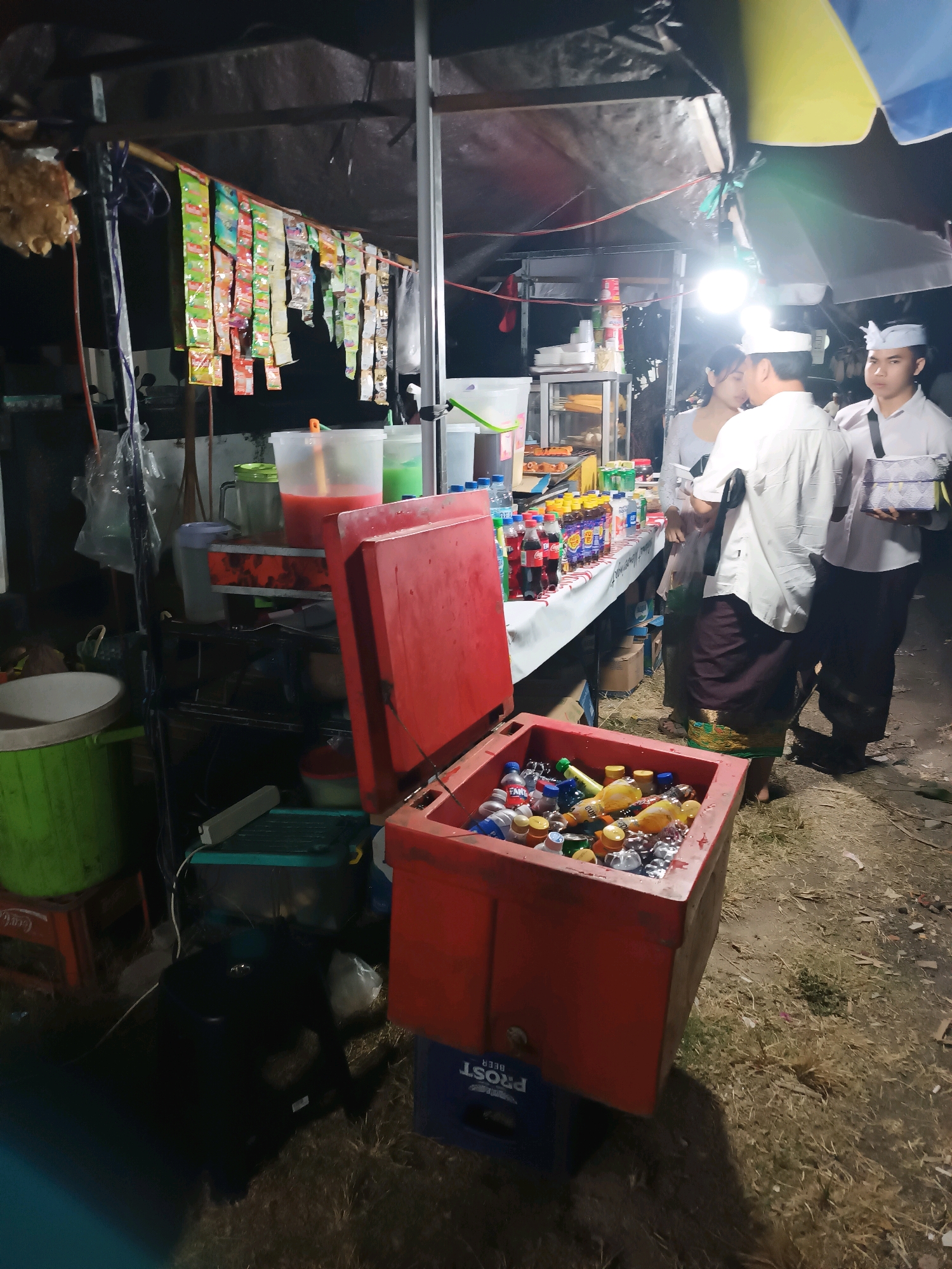 The image depicts a street-side food and beverage stall, likely in the city of Kediri, Indonesia, at night. The stall is set up under a makeshift canopy with bright artificial lighting illuminating the scene. In the foreground, a large red cooler is filled with an assortment of bottled drinks, many of which are submerged in ice. Several bottles are visible, including brands like "Fanta" and "Sprite," indicating a selection of soft drinks.
Behind the cooler, a table is laden with more beverages and what appear to be snacks or food items. Bottles of various sizes and colors are neatly arranged, along with some stacked cups. A large transparent container holds a red liquid, possibly a fruit juice or beverage concentrate. To the left, a green bucket and a beige container are visible.
Three individuals are present at the stall. Two men, dressed in white shirts and traditional dark-colored sarongs or skirts, appear to be attending to or interacting with customers. One of the men is holding a stack of what might be packaged goods or promotional items. A woman, wearing a light-colored top and a dark sarong, is also at the stall, possibly a customer or another vendor. The ground beneath the stall is covered in dry grass and some debris, typical of an outdoor setting. The overall atmosphere suggests a busy night market or street vendor operation.