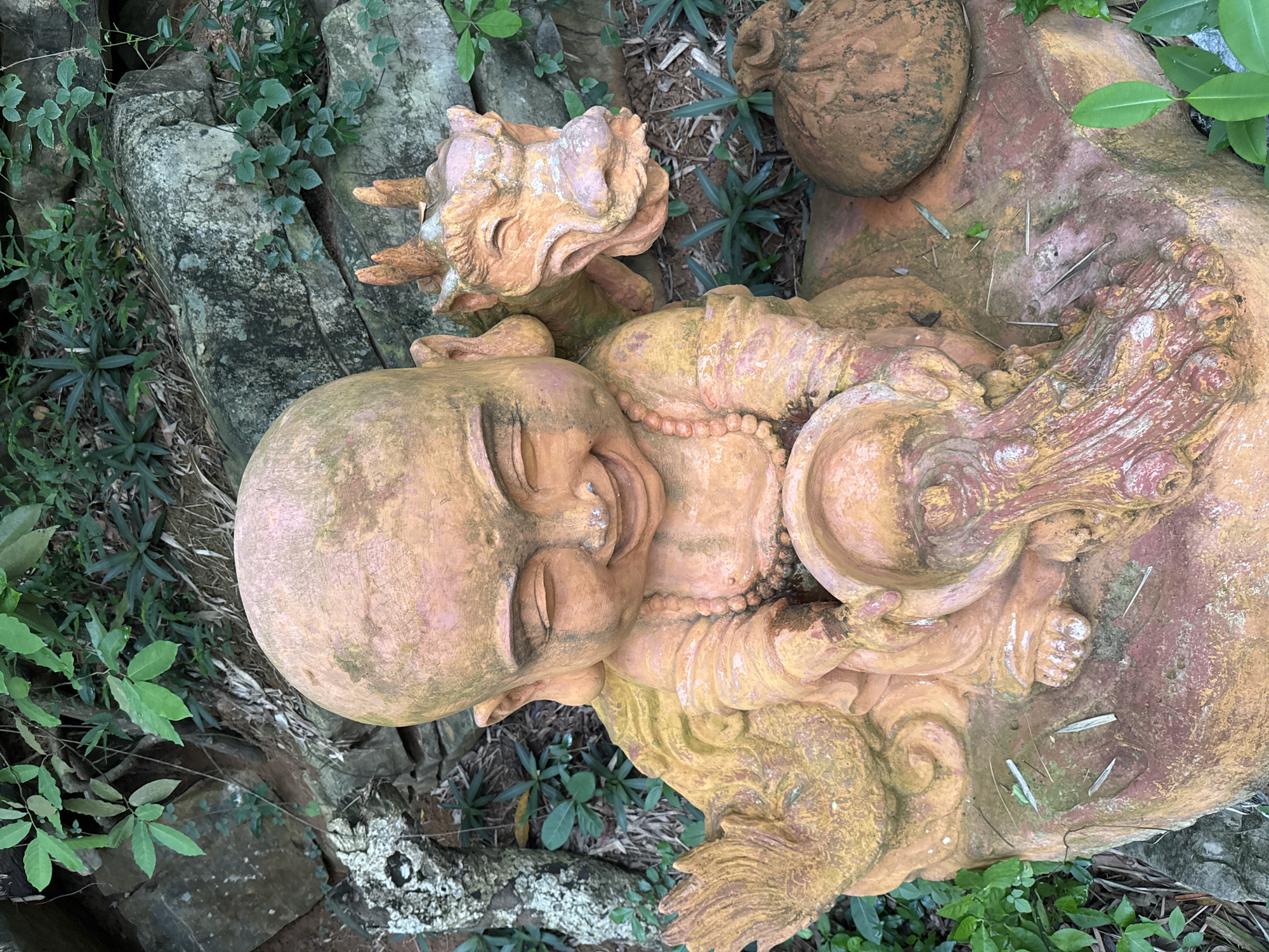 The image depicts a terracotta statue of a smiling child monk, seated with a small dragon perched on its right shoulder. The monk is holding a bowl from which a stream of water appears to be flowing. To the right of the monk, there is a sack-like object. The statue is situated outdoors, surrounded by rocks and lush green foliage, suggesting a garden or natural setting. The statue itself is weathered, with moss and dirt visible on its surface. The scene appears to be located in the city of Tây Hoa Lư Ward, Vietnam, a region known for its historical sites and natural beauty.