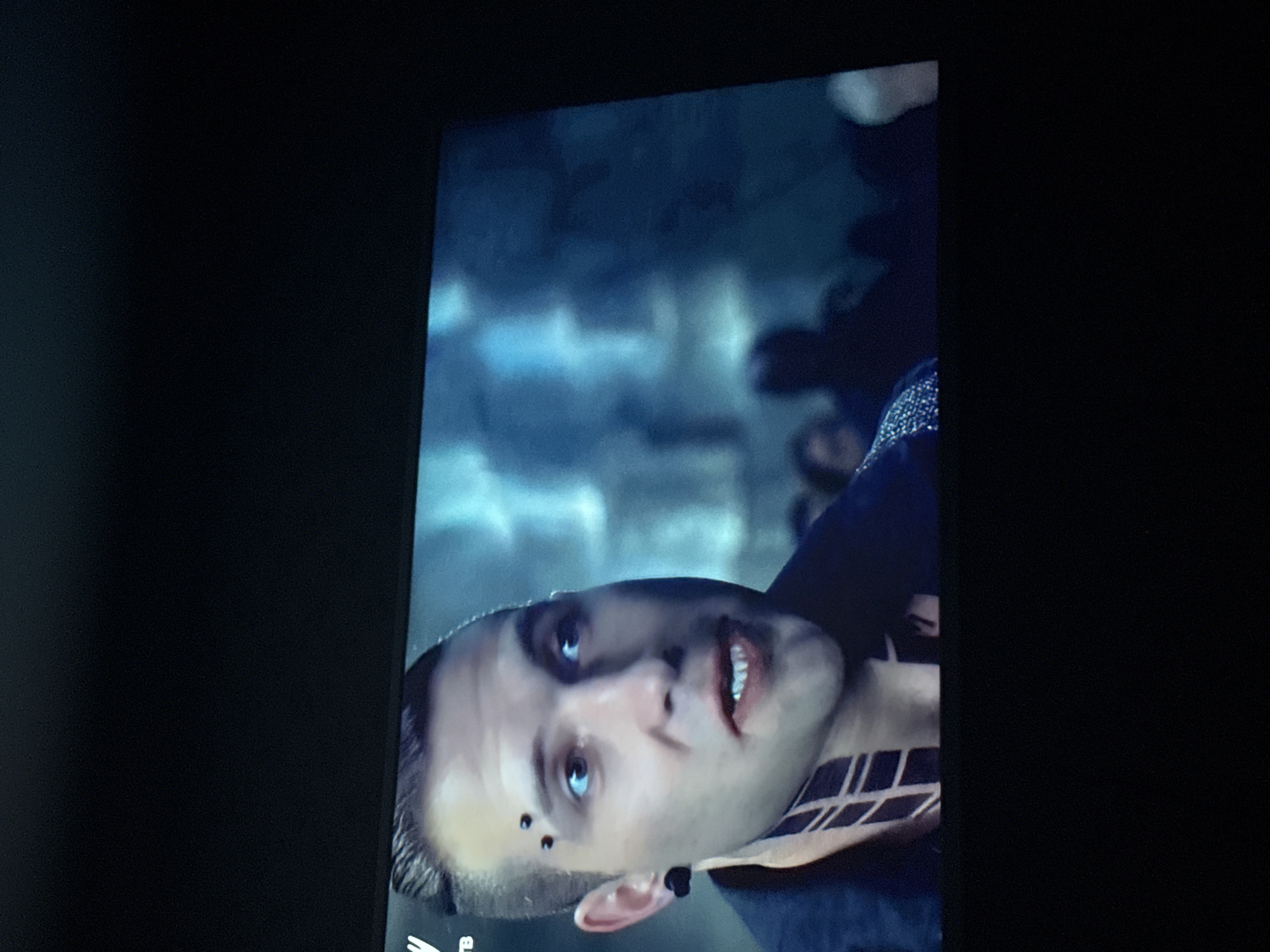 The image shows a close-up of a man's face. He has pale skin, blue eyes, and a dark piercing above his left eyebrow. There is a tattoo of dark geometric shapes on his neck, visible just below his jawline. His hair is short and styled back. The background is blurred, showing indistinct figures and structures, suggesting an urban setting.
The city of Mytishchi, Russia, is not discernible from the details provided in this image.