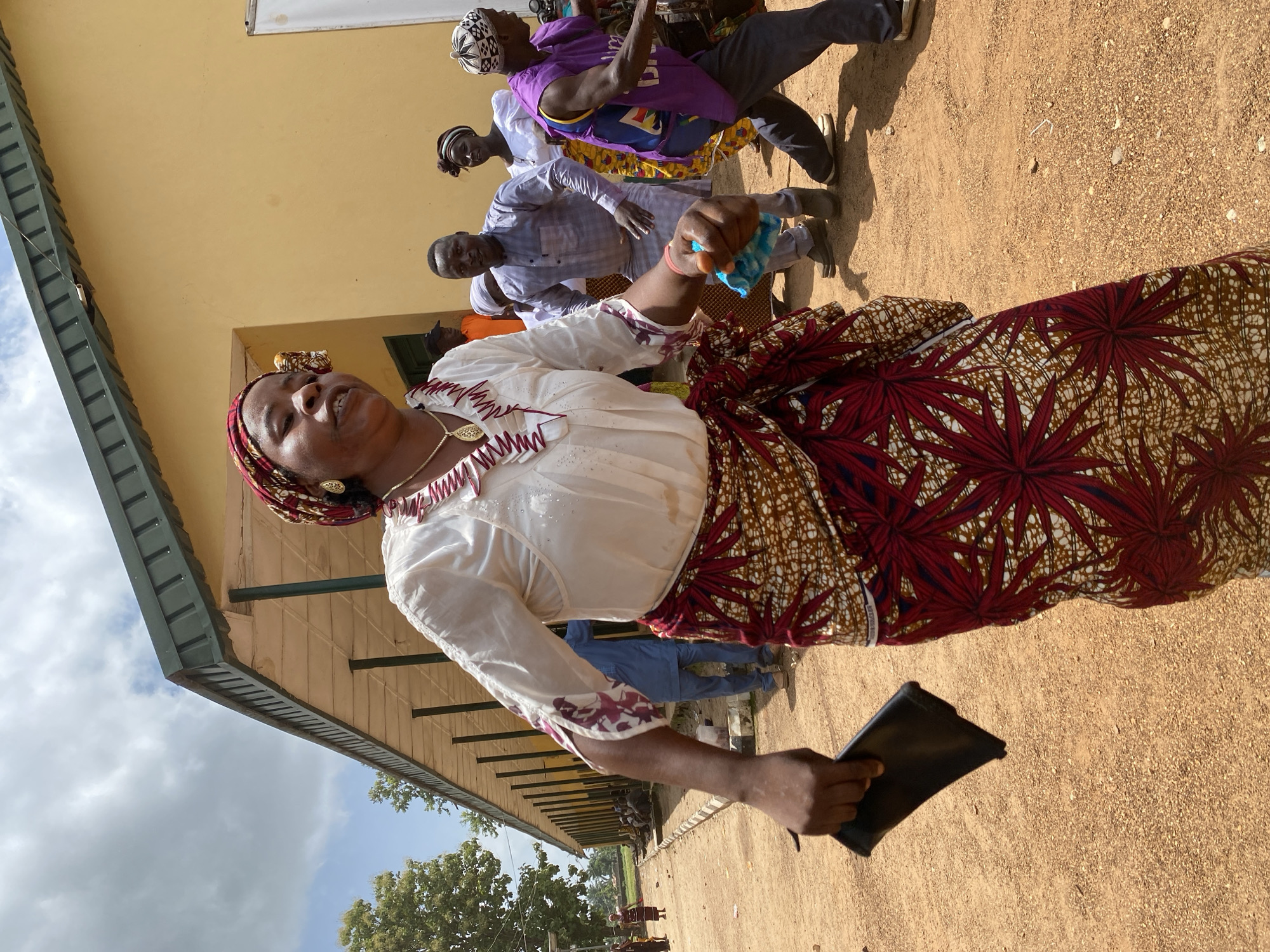 In Maigizo, Nigeria, an African woman stands on a reddish-brown, gravelly ground, possibly a schoolyard or public space. She wears a patterned headwrap, a white blouse with red trim, and a red and brown patterned skirt. She is holding a black tablet or book in one hand and a crumpled blue item in the other. Several other African people are behind her; some are dancing. A long, single-story building with a light-yellow wall and a dark-green roof runs along the left edge of the image. Trees are visible in the distance. The sky is cloudy.