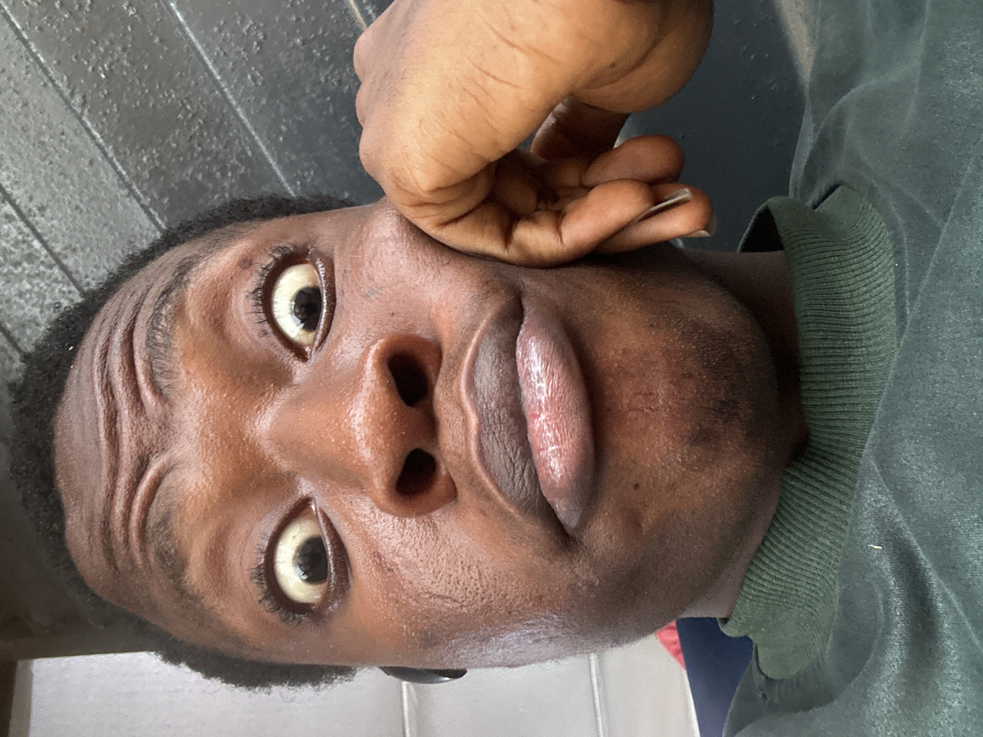 A close-up, upward-facing view of a dark-skinned person with short, dark hair. Their eyes are wide open, and they are making a pursed-lip expression. A hand is near their face, possibly touching the side of their head. They are wearing a dark green, collared shirt. The background appears to be a dull, dark grey surface. The location is Unknown, Unknown.