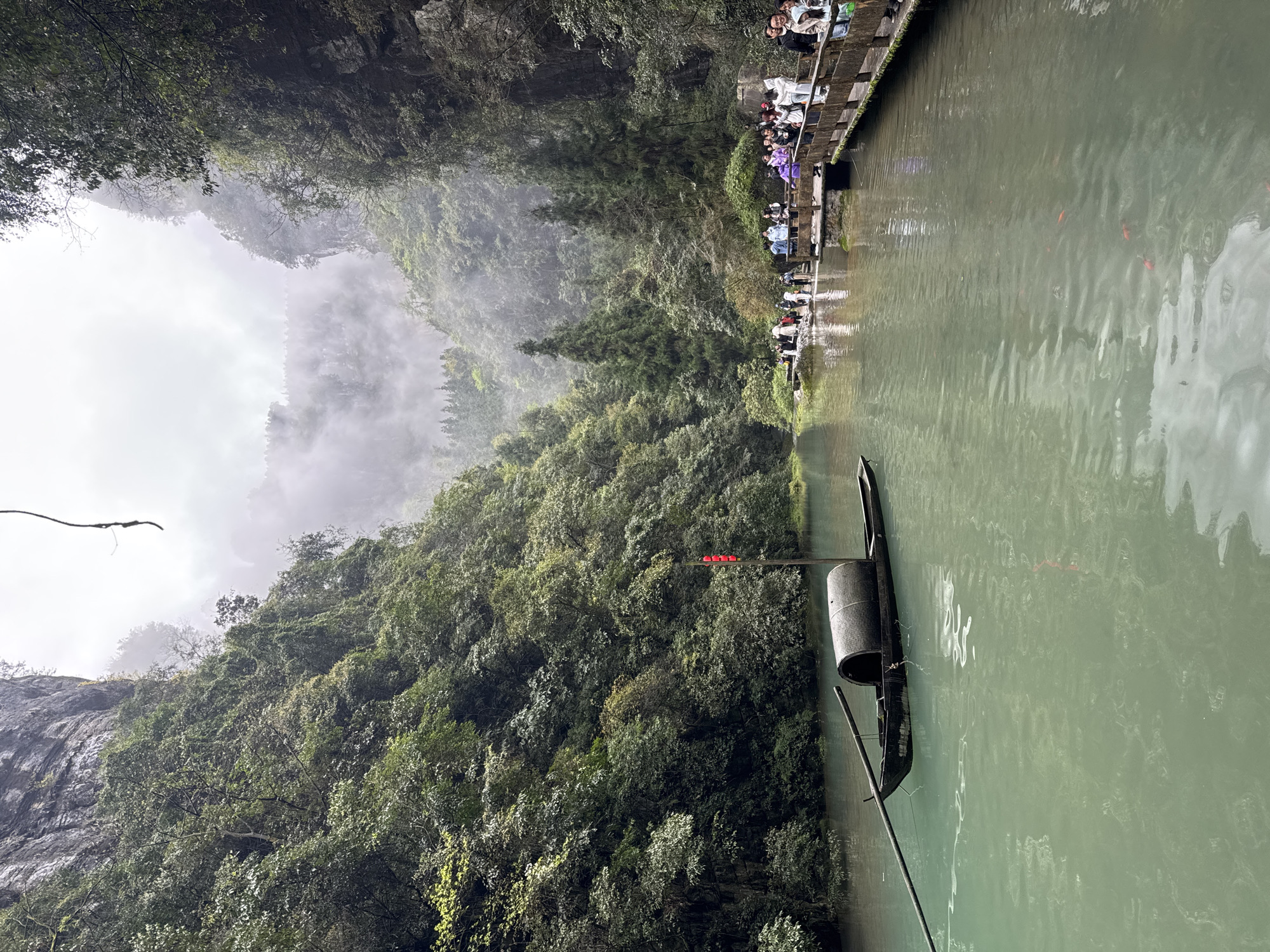 The image depicts a boat on a river in the Wulong District, Chongqing, China. The river is bordered by dense green vegetation on one side and a stone walkway crowded with people on the other. Mountains are visible in the background, obscured by mist or fog. The boat is long and narrow with a dark hull and a cylindrical container on board. The water is a muted green color with ripples and possible fish visible.