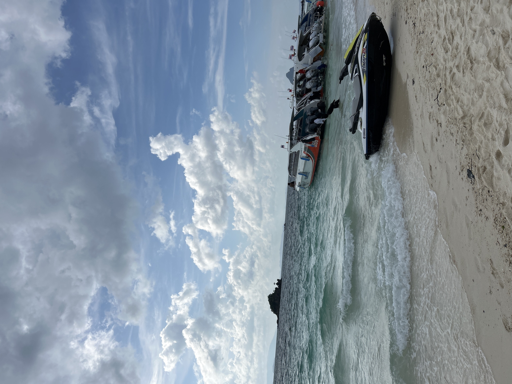 The image shows a tropical beach scene. In the foreground, a jet ski is parked on the sand, with the waves lapping at its hull. The sand is light in color and shows footprints. Further out, a line of speedboats is anchored in the turquoise water. The boats are of various colors and designs, some with multiple outboard motors. In the distance, a small, rocky island is visible. The sky above is partly cloudy, with patches of blue sky showing through. The overall impression is one of a popular tourist destination, likely in a coastal area of Thailand, given the boats and the island formation. The specific location is the beach of Phru Nai, Thailand.