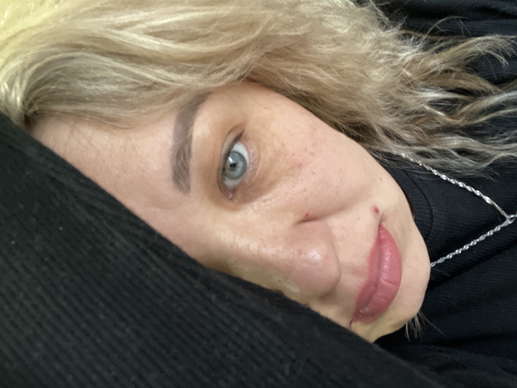 A close-up, slightly off-center image shows a woman with light blonde, curly hair partially covering her face. Her skin is light with visible pores and some red spots. She has one visible eye, which is a light blue color. Her lips are full and appear to have lipstick or lip gloss applied. She is partially covered by a dark, possibly black, garment, which also serves as the background in some areas. A silver chain necklace is visible on her neck. There's no information to determine the location in the city of Chaikovsky, Russia.