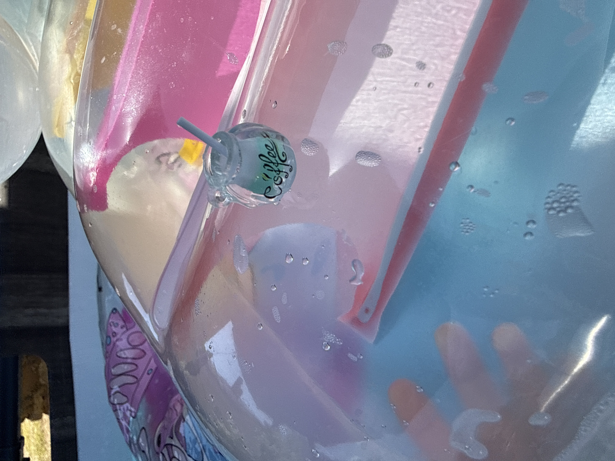 The image depicts a close-up view of a clear inflatable object, possibly a pool float or inflatable toy, filled with a light blue liquid and containing air bubbles. A hand, with fingers partially visible and submerged in the blue liquid, is pressing against the inside surface of the inflatable. A small glass jar with a straw is attached to the exterior of the inflatable, and the word "coffee" is written in cursive on the side of the jar. The background shows other colored inflatable objects, including one with pink and yellow sections, and a patterned design in pink and purple. The scene appears to be outdoors, possibly in a yard or park setting, with a blurred background that suggests natural elements and structures. There is no identifiable information indicating a specific location in the city of Gara Oreshets, Bulgaria.
