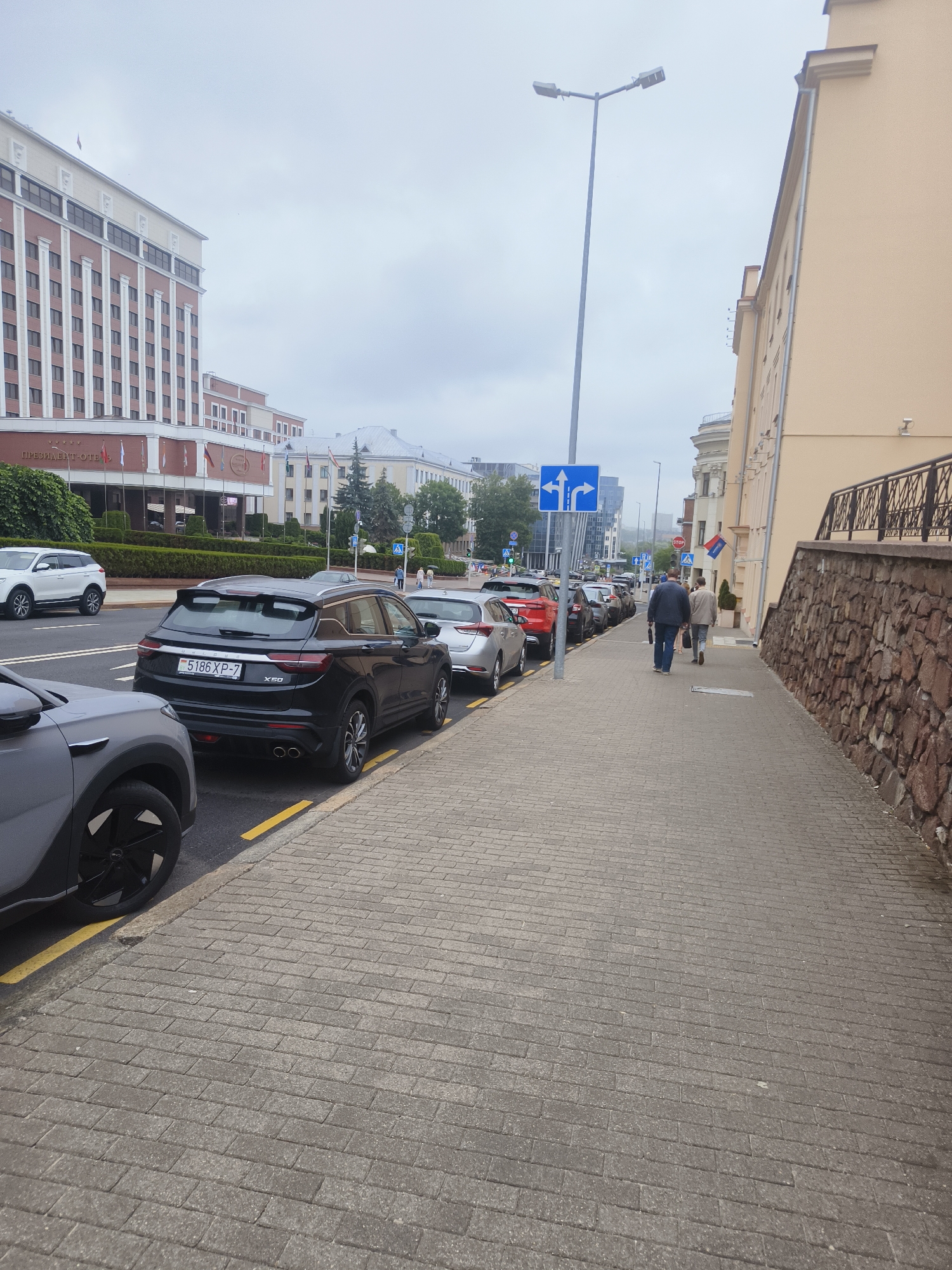 The image depicts a street scene in Minsk, Belarus, likely near a prominent building. On the left, a large, modern hotel with a red brick facade and white columns is visible, identified by the sign "ПРЕЗИДЕНТ-ОТЕЛЬ" (President-Hotel) with Russian Cyrillic lettering. A row of parked cars lines the street in front of the hotel, including a black Geely Coolray with a license plate reading "5186 XP-7". A grey SUV is parked in the foreground on the left.
The sidewalk to the right is paved with grey brickwork. Two men are walking away from the camera on this sidewalk. One man wears a dark jacket and blue jeans, and the other wears a light-colored jacket and dark trousers. They are walking towards the right side of the image.
Further down the street, more cars are parked. A traffic light with directional arrows indicates turning lanes. A stone wall borders the sidewalk on the right, with a metal railing on top. The sky is overcast.