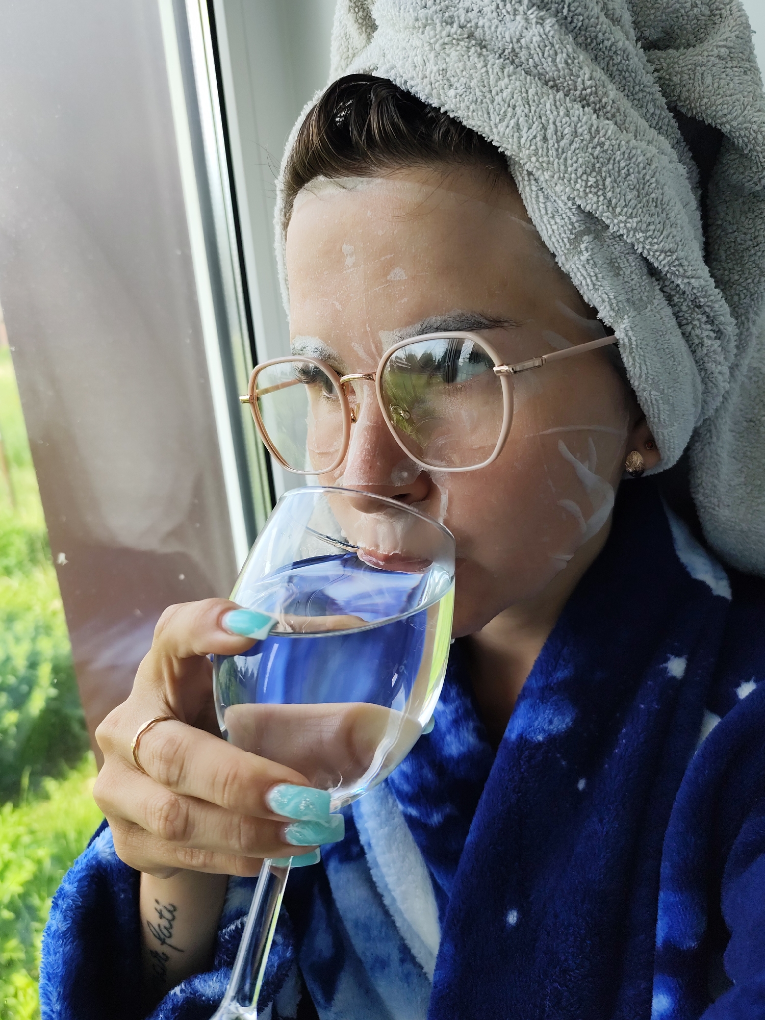 A woman is drinking from a wine glass filled with water. She is wearing a gray towel wrapped around her head, a blue robe with white star patterns, and gold-rimmed glasses. There is a white face mask applied to her face. Her nails are painted light blue with a textured design, and she has a gold ring on her finger. A tattoo is visible on her wrist. The background shows a window with green vegetation outside. The location in the city of Фоки, Russia is not evident.