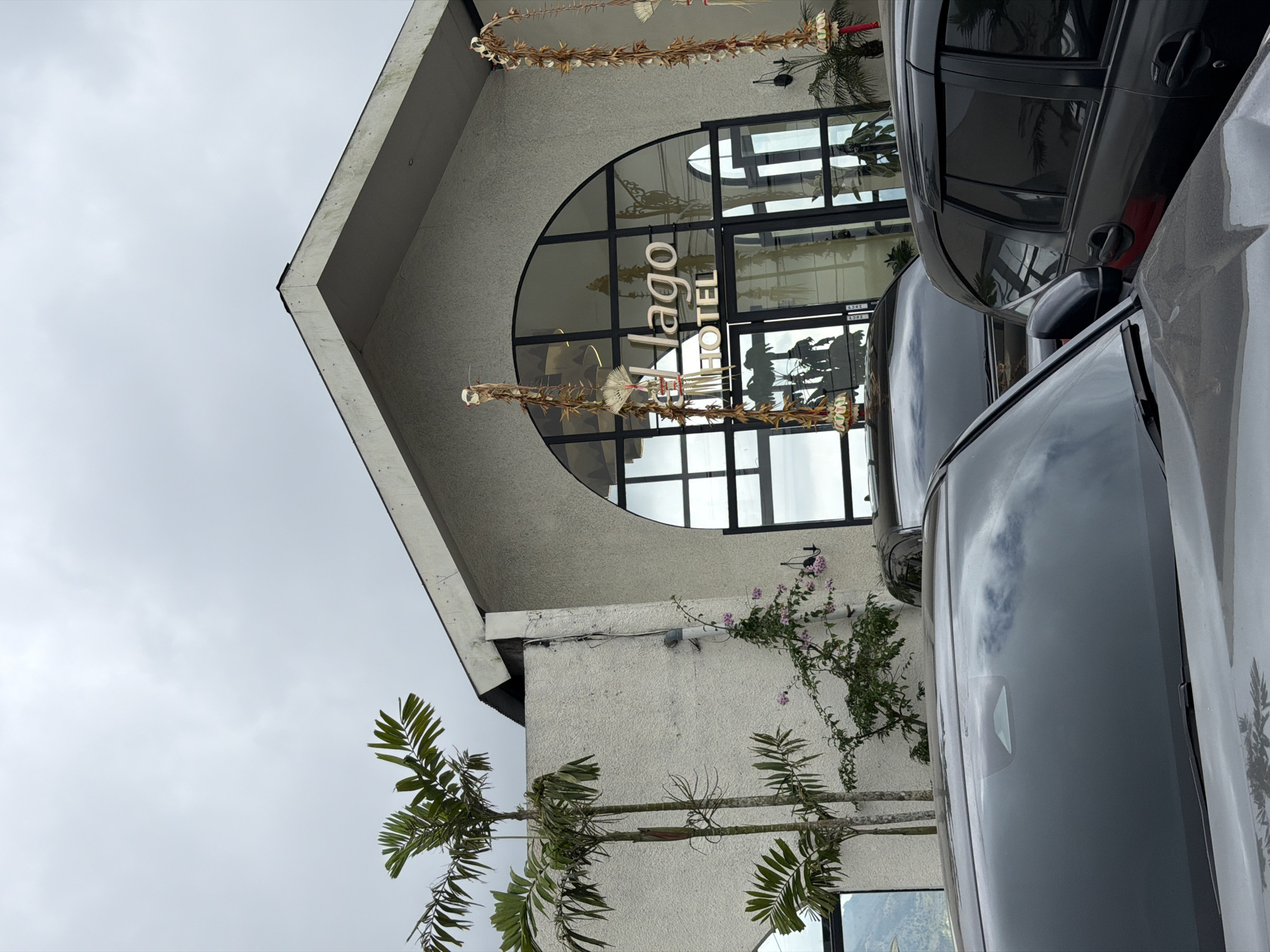 The image depicts the entrance of the Lago Hotel in Kintamani, Indonesia, on an overcast day. The hotel features a modern architectural style with a textured white facade and a large arched glass entryway. Decorative Balinese offerings, known as "canang sari," are hung on either side of the entrance. In front of the hotel, several dark-colored cars are parked, partially obscuring the building's lower section. Two palm trees and some flowering vines are visible to the left of the entrance, adding a touch of tropical greenery to the scene. The overall impression is one of a serene, slightly humid atmosphere typical of the Kintamani region.