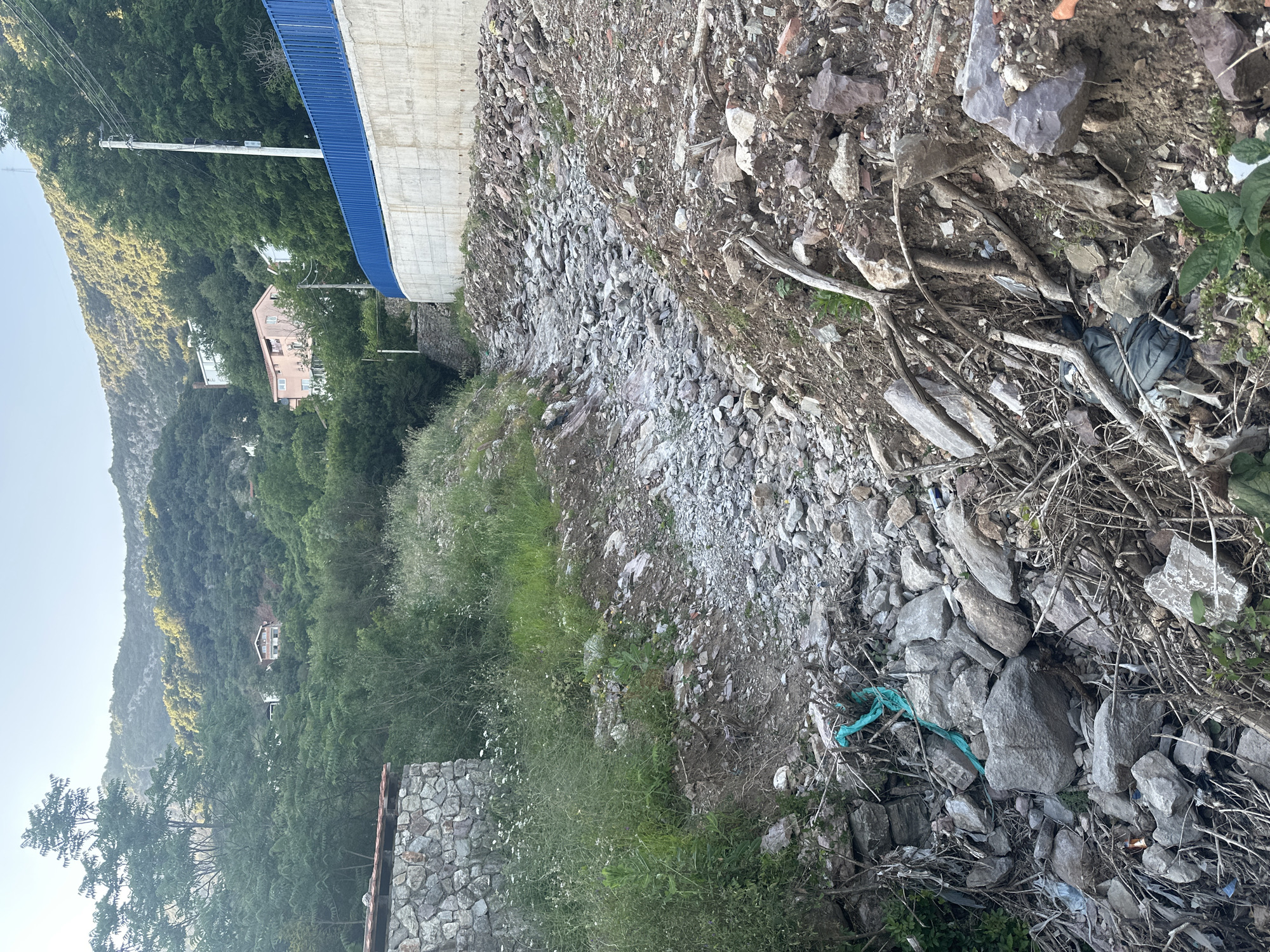 The image depicts a rugged, sloped terrain consisting of dirt, rocks, and scattered vegetation. A blue-trimmed concrete structure is visible on the left side of the frame. In the background, there is a mountainous landscape with houses scattered amongst the trees. The location is likely a hillside on the outskirts of Budva, Montenegro. Scattered trash is visible in the foreground. A blue rope lies among the rocks.