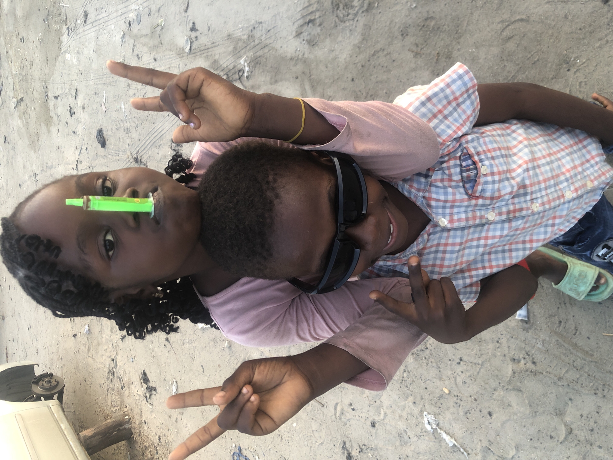 In Ado, Nigeria, a young girl stands behind a younger boy, her arms around his shoulders. She is holding a green syringe vertically on her nose and making a peace sign with her left hand. The boy, wearing sunglasses and a checkered shirt, also makes a peace sign with his right hand. They are standing on sandy ground near what appears to be a disassembled car. The overall scene suggests a casual, playful moment outdoors.