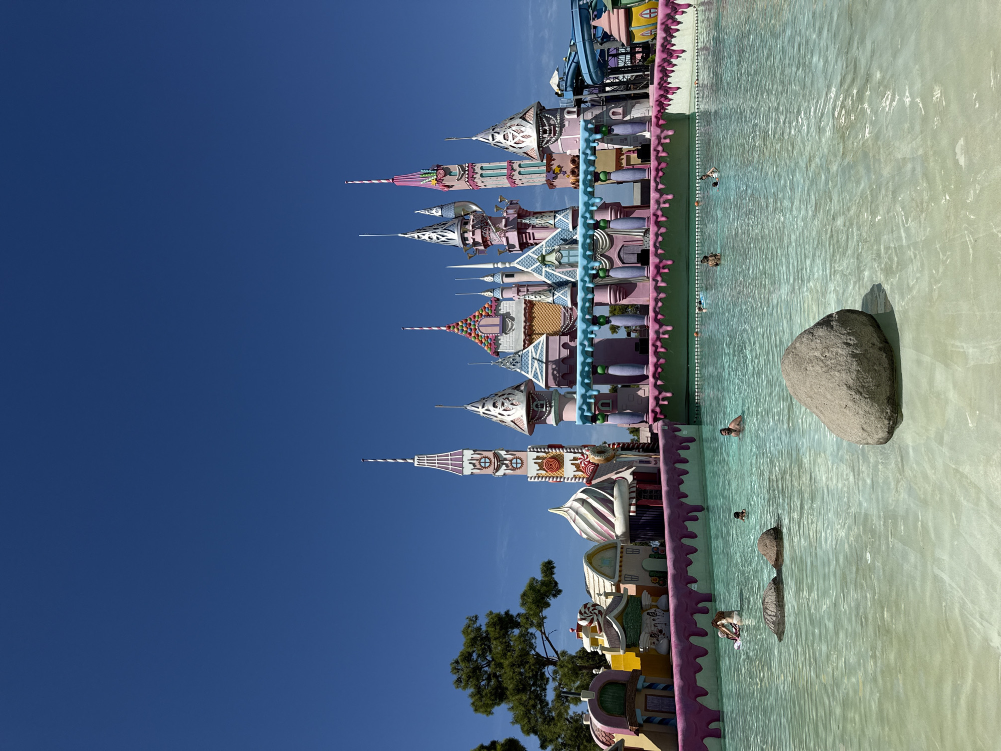 The image shows a bright, sunny day at a water park in Bodrum, Turkey. In the foreground, a shallow, clear blue pool has several large rocks partially submerged. A few people are swimming and relaxing in the water. In the background, a whimsical, candy-themed structure resembling a castle dominates the scene. The castle is adorned with pastel colors, candy-like decorations, and multiple spires. Adjacent to the castle is a water slide. The sky is a clear, vibrant blue.