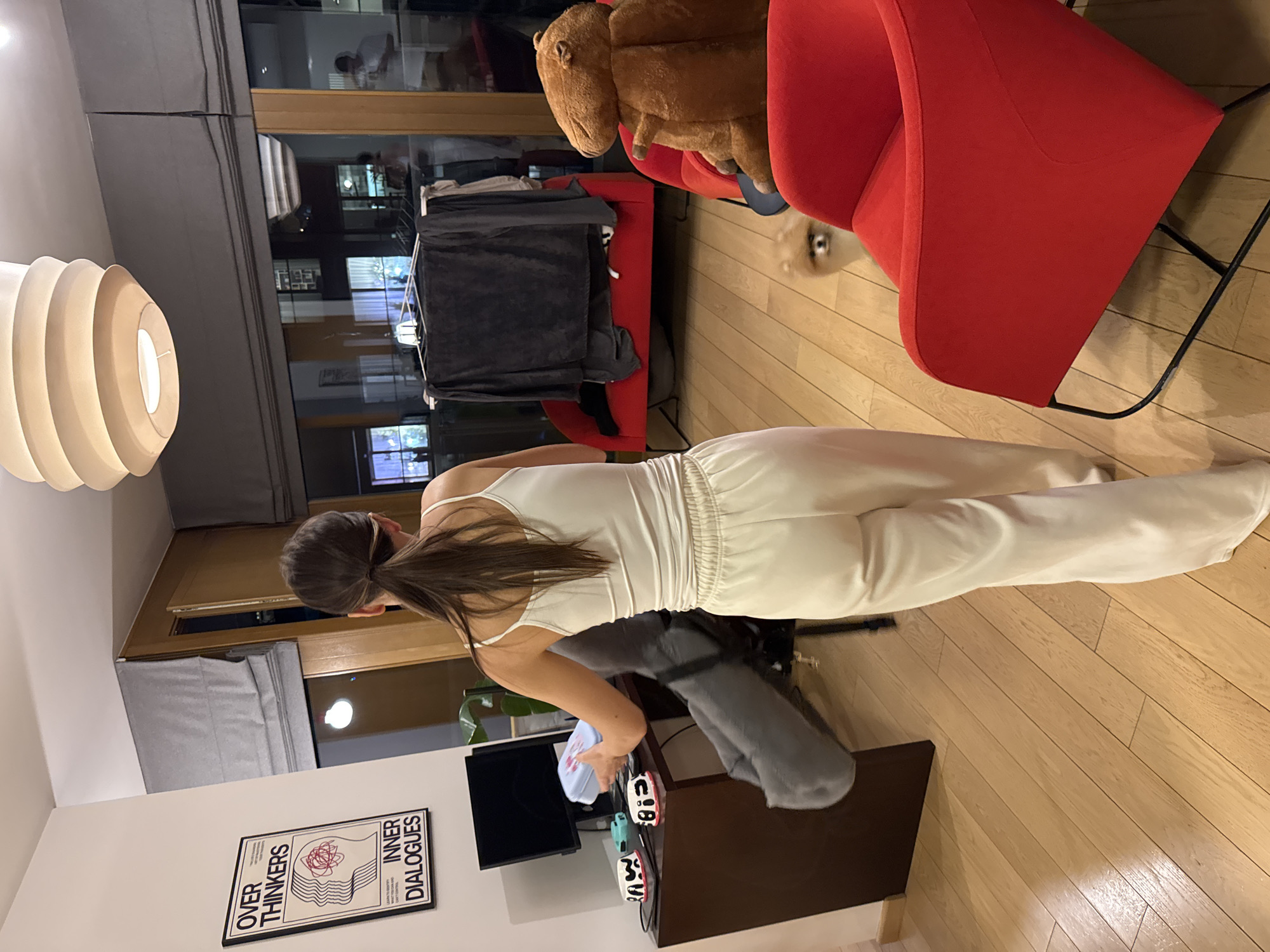 A woman with long dark hair in a ponytail, wearing a white spaghetti-strap top and white pants, is standing in a room. Behind her, there's a dark brown desk with a computer monitor, a gray garment, and a couple of decorative figurines. On the wall next to the desk is a framed poster with the text "OVER THINKERS INNER DIALOGUES." In the background, there's a red armchair, a stuffed animal on a red bench, and a clothes drying rack with garments. The room has wooden floors. Through the window, building lights are visible, suggesting an urban environment, and the location is Warsaw, Poland.