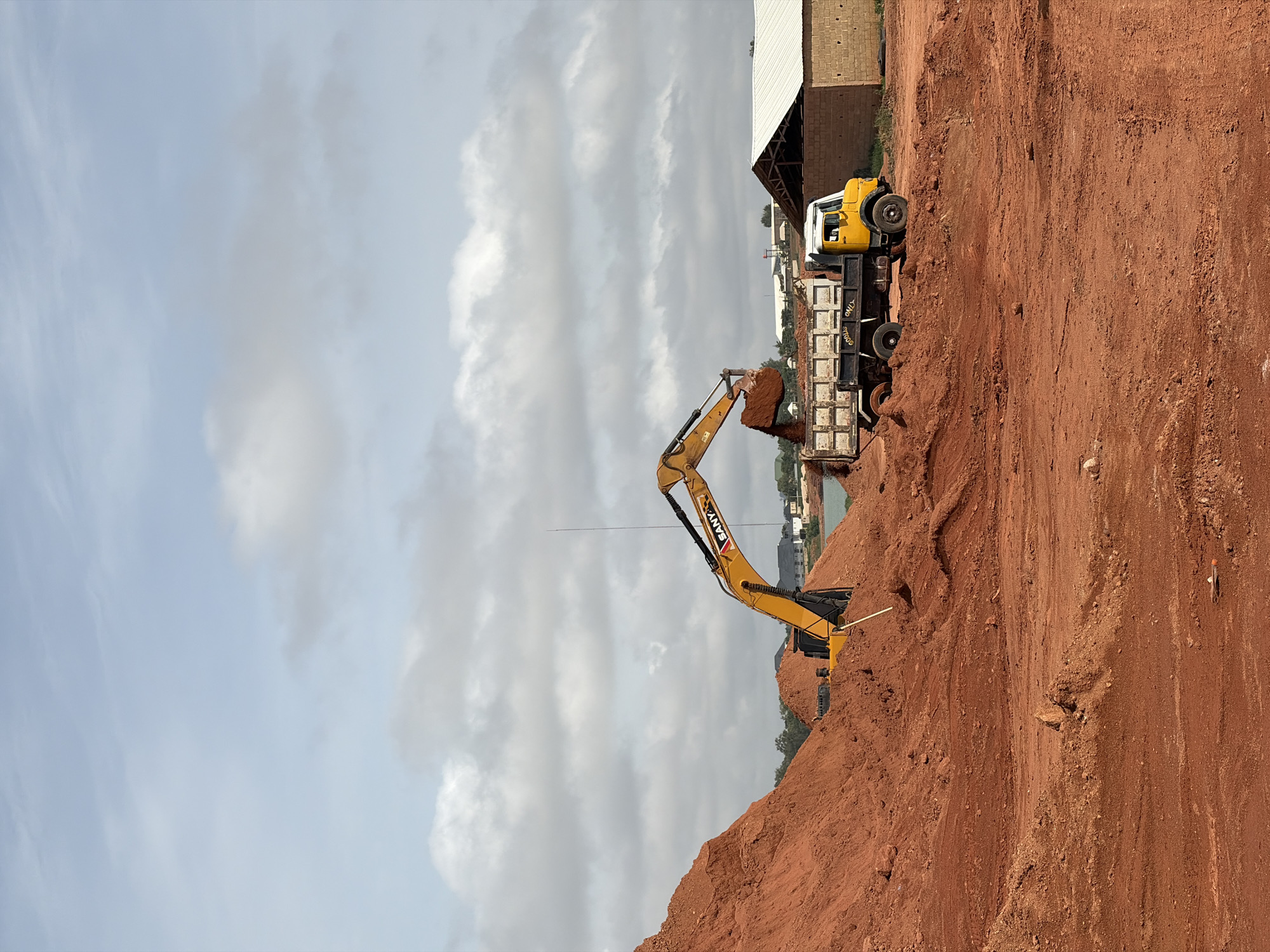 In Kwang, Nigeria, a construction site is depicted with an excavator loading dirt into the bed of a yellow dump truck. The truck is positioned to the right of the excavator, with its rear end facing the excavator's bucket, indicating an active material transfer. The ground is composed of red dirt, with large mounds of soil visible on the left and foreground. In the background, a building with a white roof and brick walls stands on the right, and a body of water can be seen behind the excavator. The sky is partly cloudy with a mix of blue and white. No people are directly visible in the scene.
